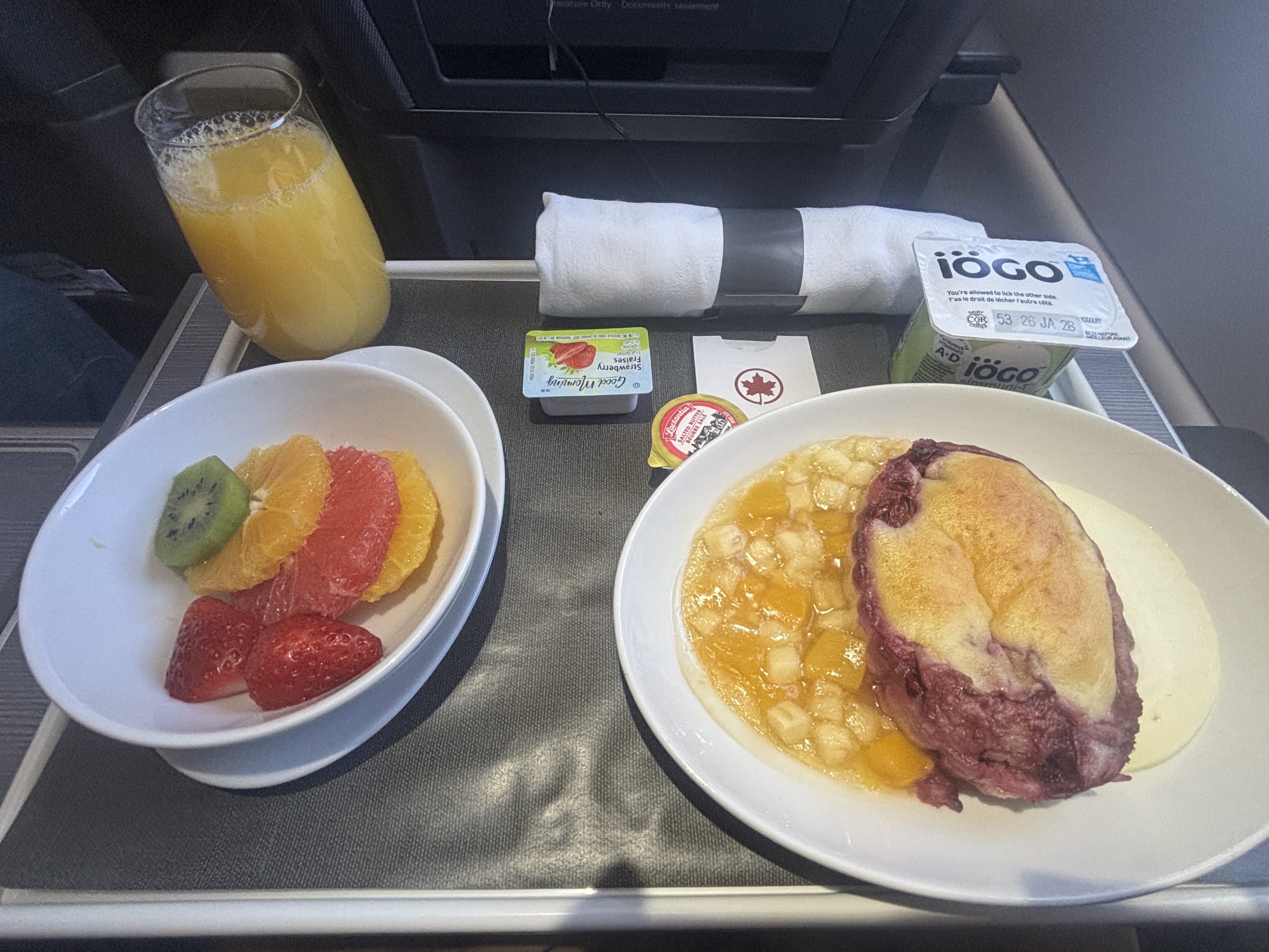 Breakfast meal tray with sliced fruit, orange juice, yogurt, and cherry clafoutis with fruit and custard.