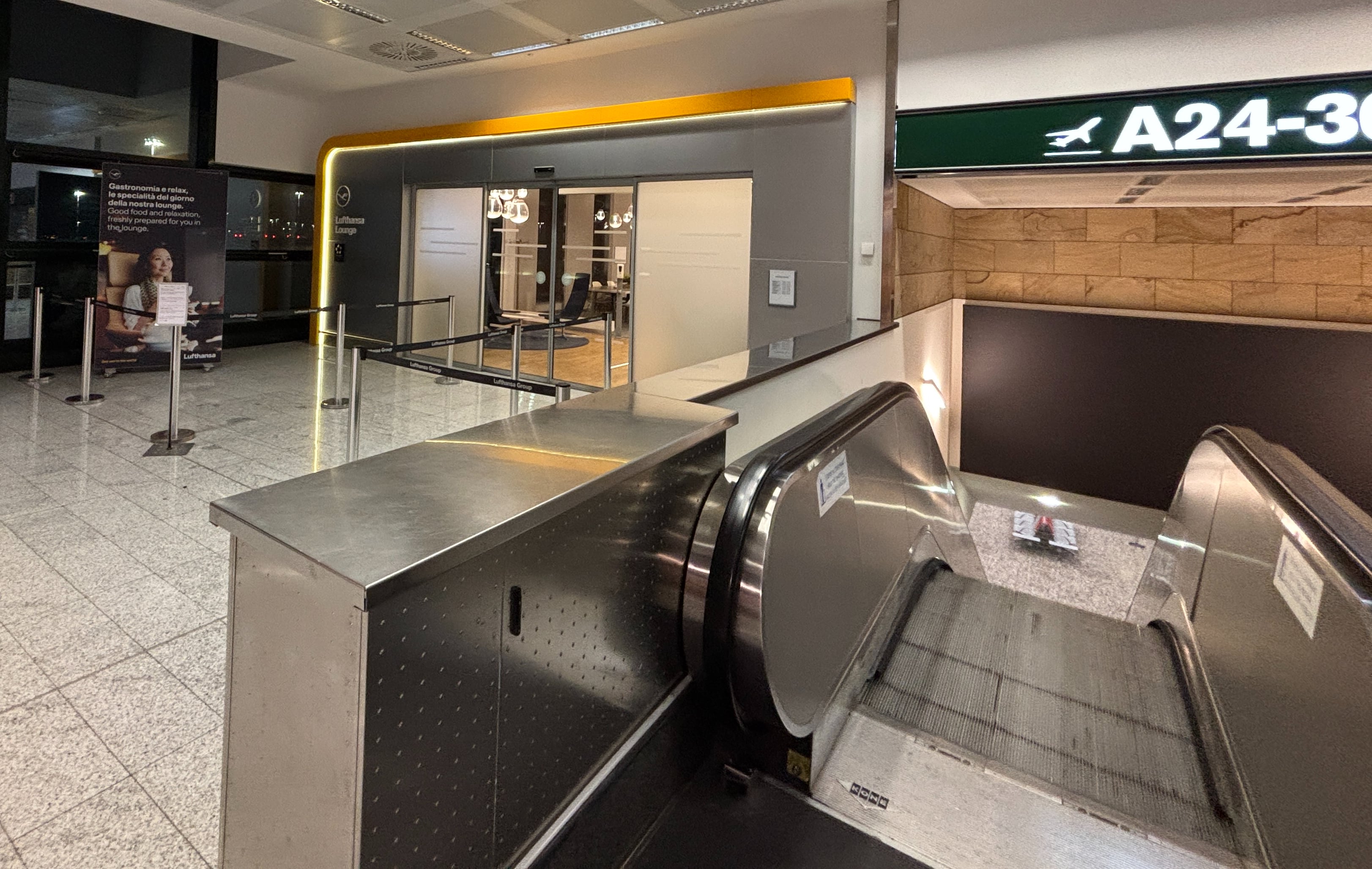 Understated entrance to the Lufthansa Lounge, a sliding glass door surrounded by backlit grey panels, next to an escalator leading downwards to the A-gates at Milan Malpensa Airport.