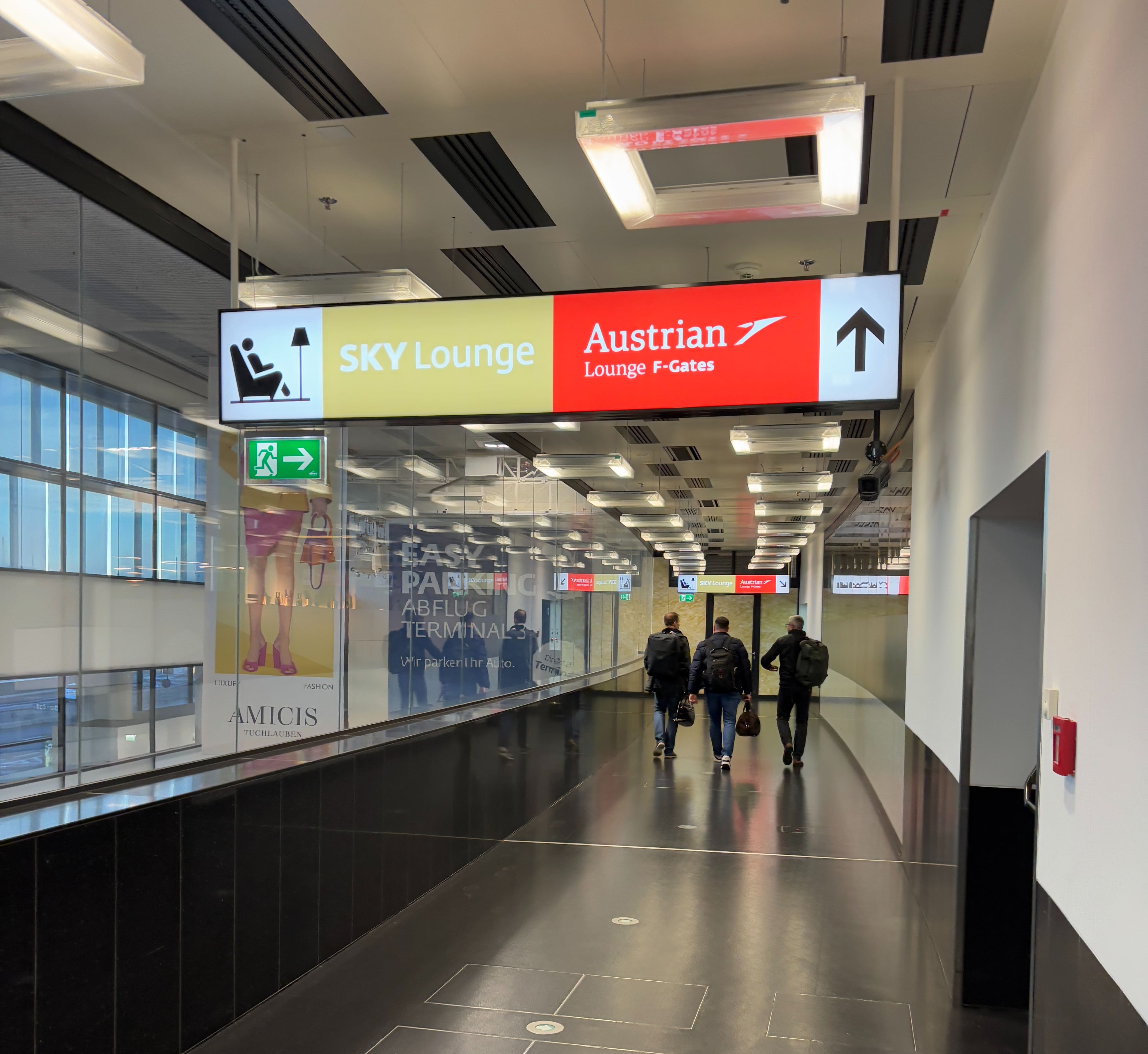 Corridor with signage indicating the entrances to the SKY Lounge and Austrian Lounge are ahead, with the public terminal areas visible out of glass on the left.