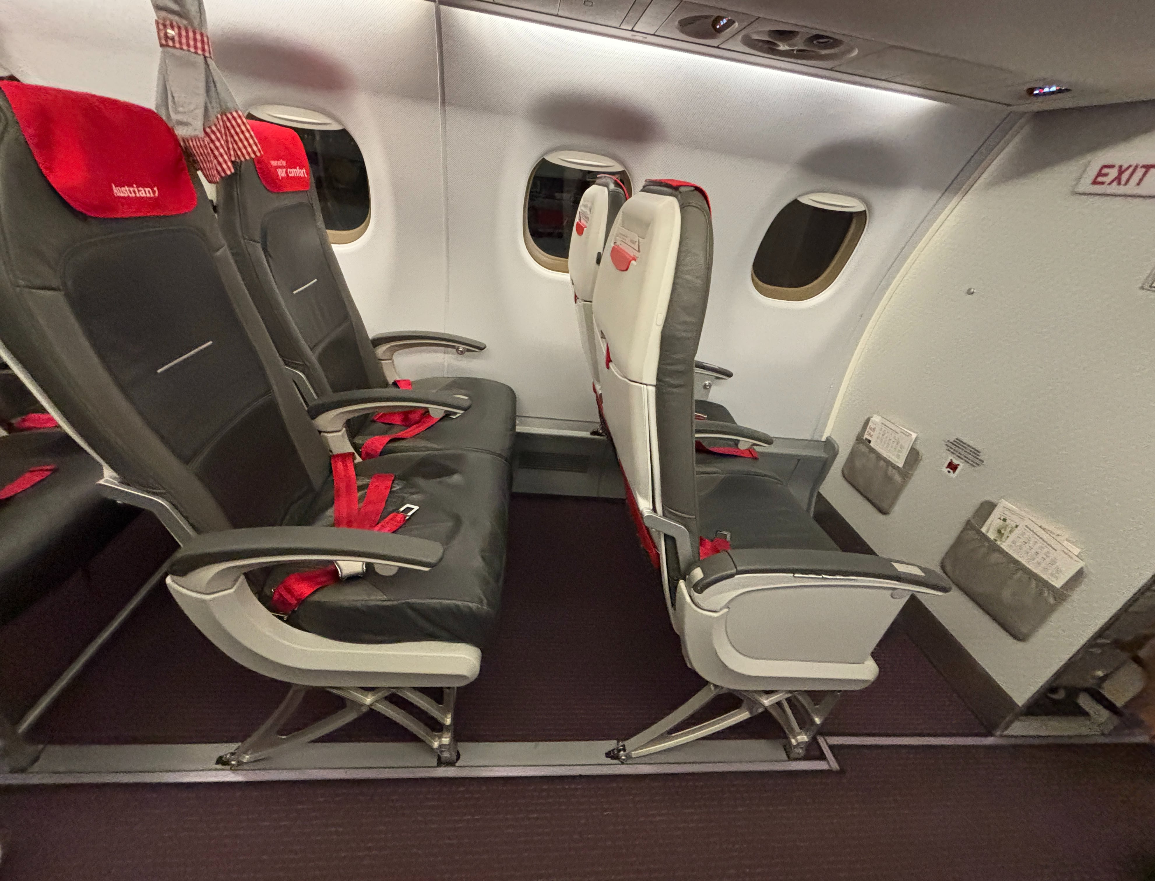 Two rows of two slimline seats with grey material and red headrests, with red-and-white checkered curtain visible dividing economy from business class.