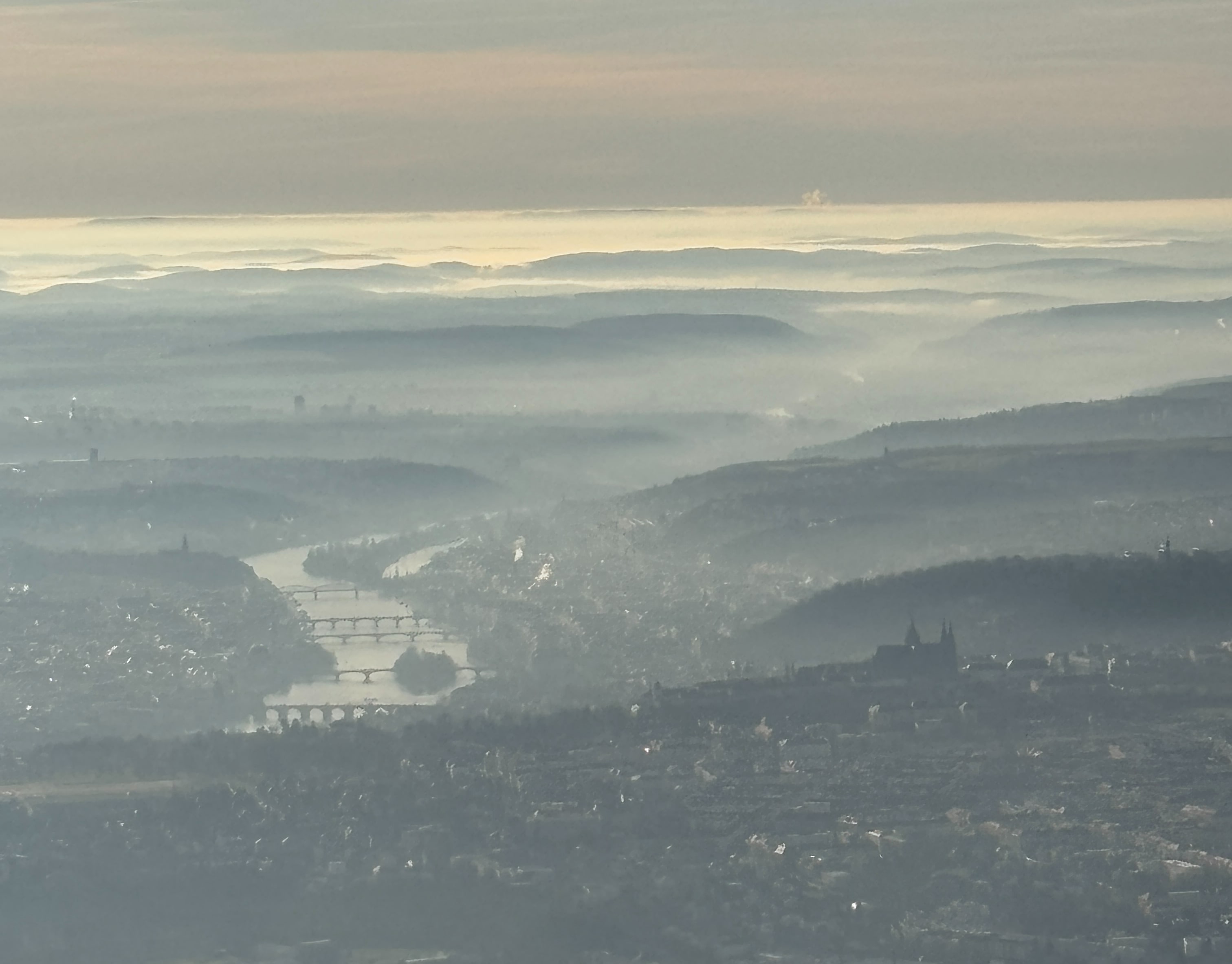 View of Prague from above, slightly obscured by fog: Vltava River and five bridges seen on the left, Prague Castle on the right.