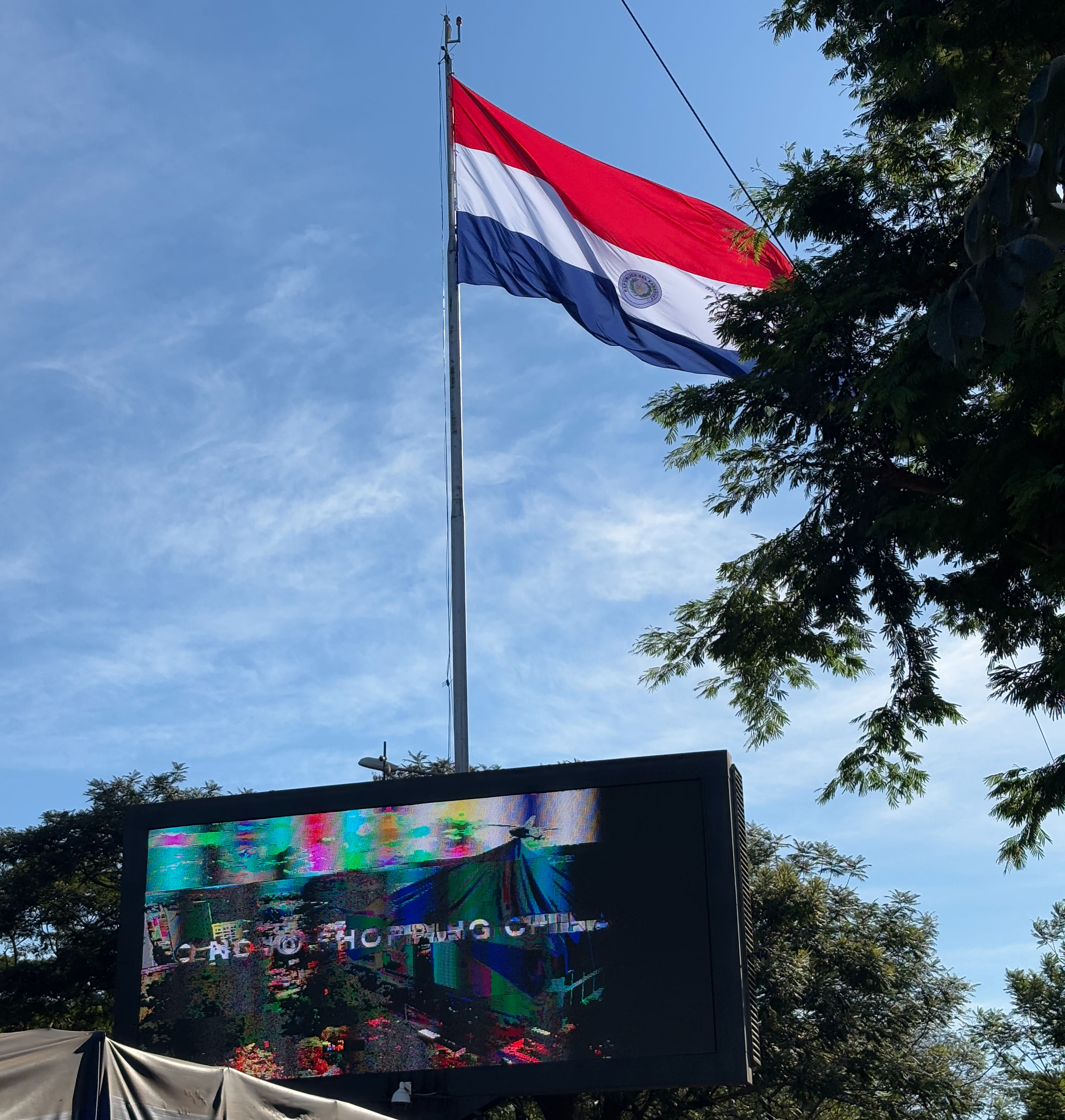 Large Paraguayan flag backlit by the sun, above a digital advertising screen transitioning between ads.