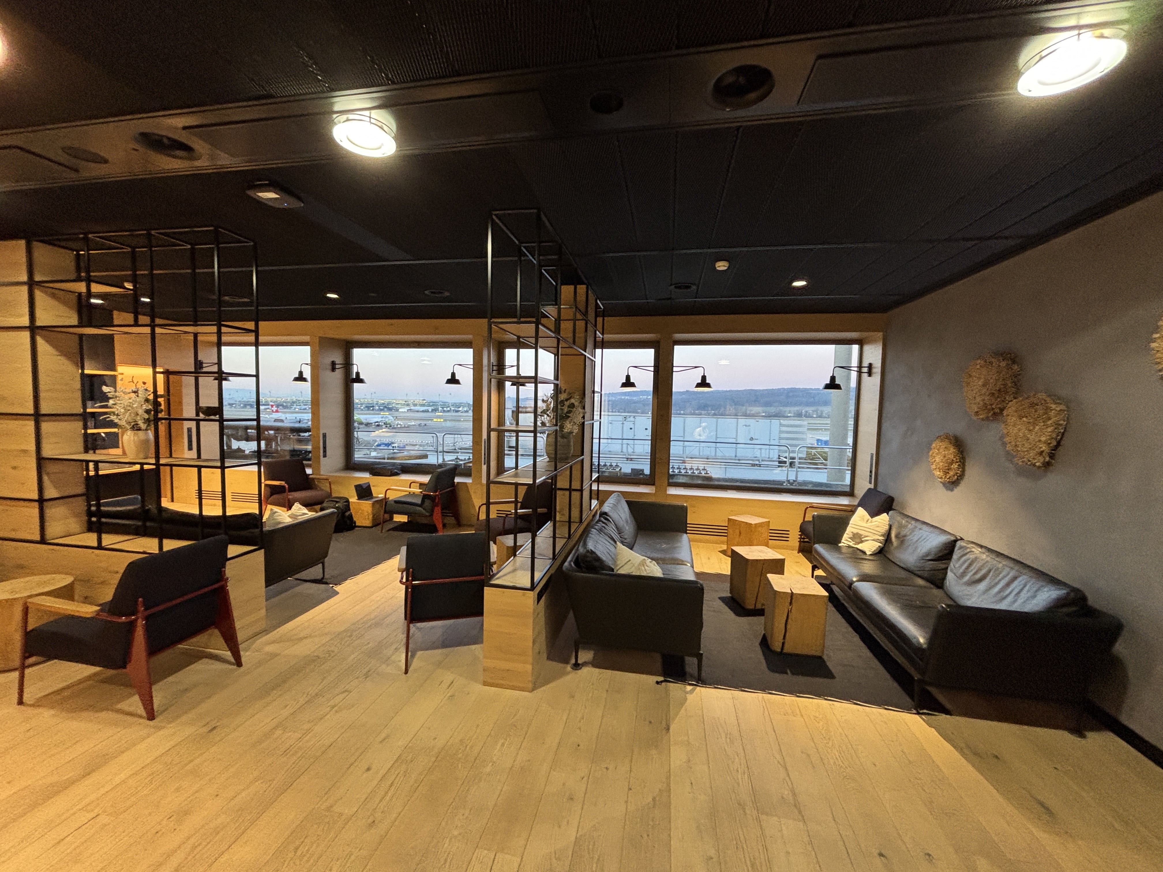 Chairs and couches arranged on a hardwood floor, with windows looking out over Zurich Airport.