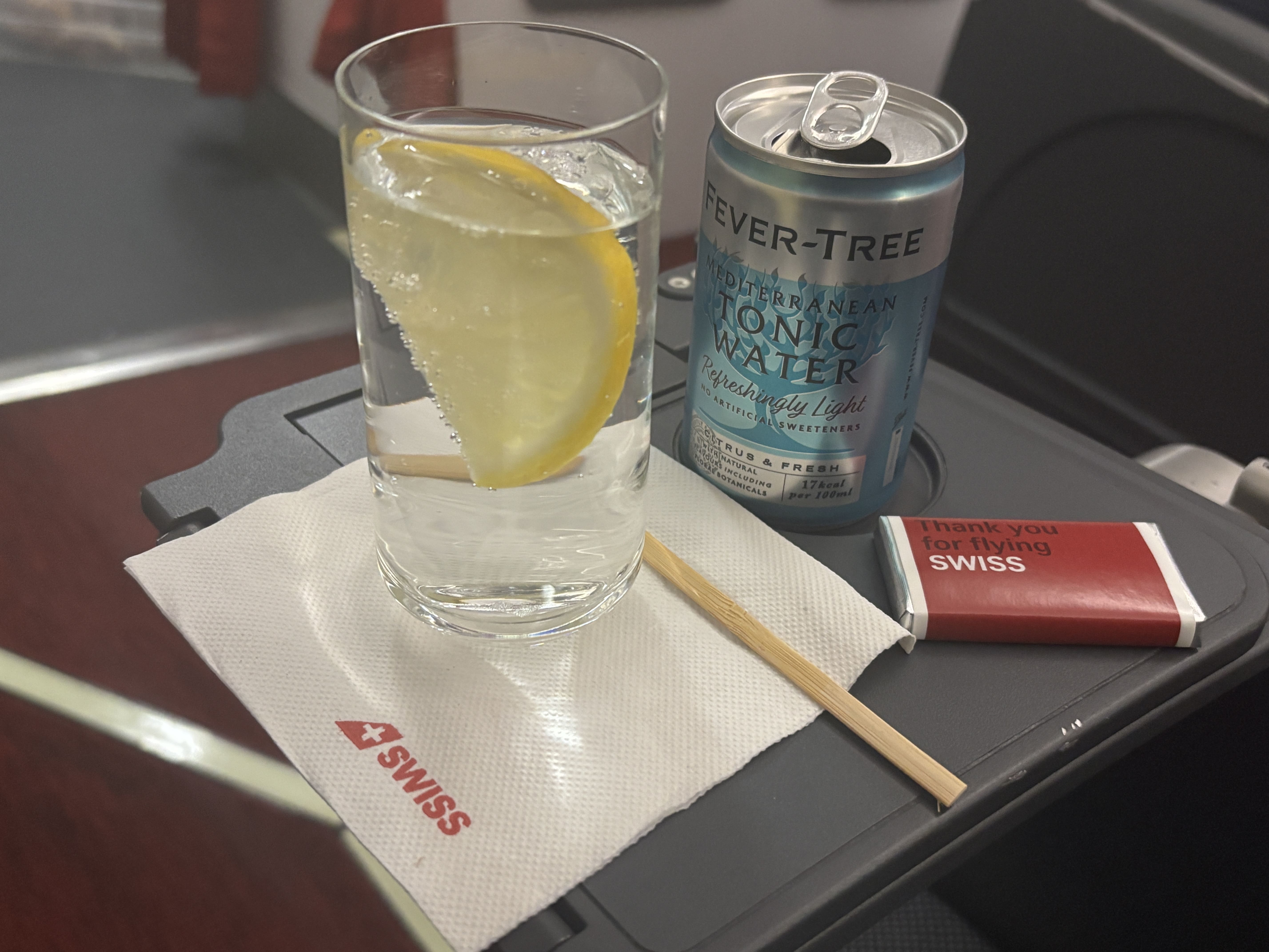 Gin and tonic with lemon slice in a clear glass on top of a half-folded tray table, with Swiss chocolate at the side.