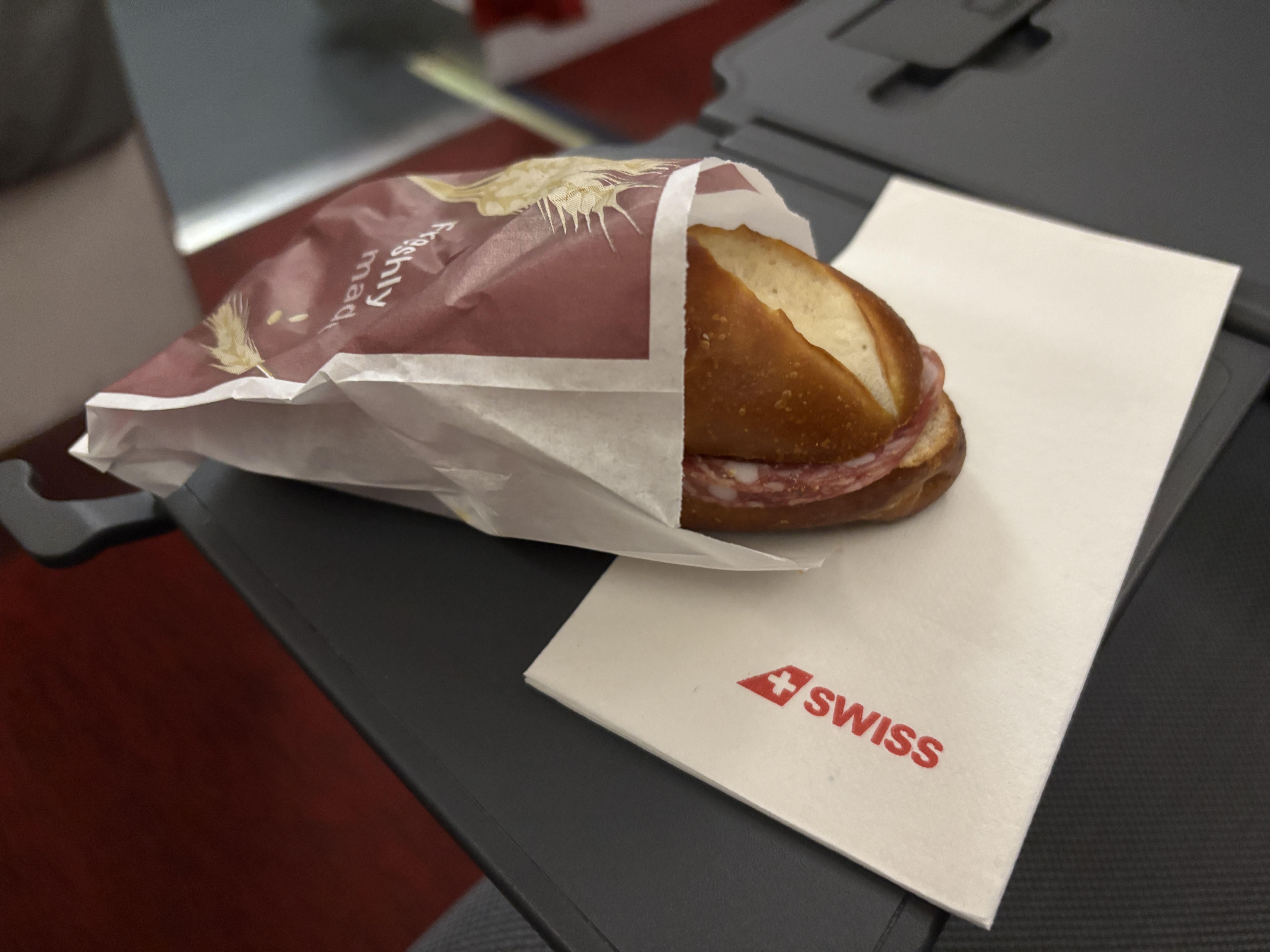 Pretzel bun containing slices of salami protruding from a paper wrapping, resting on a SWISS-branded serviette atop a tray table.