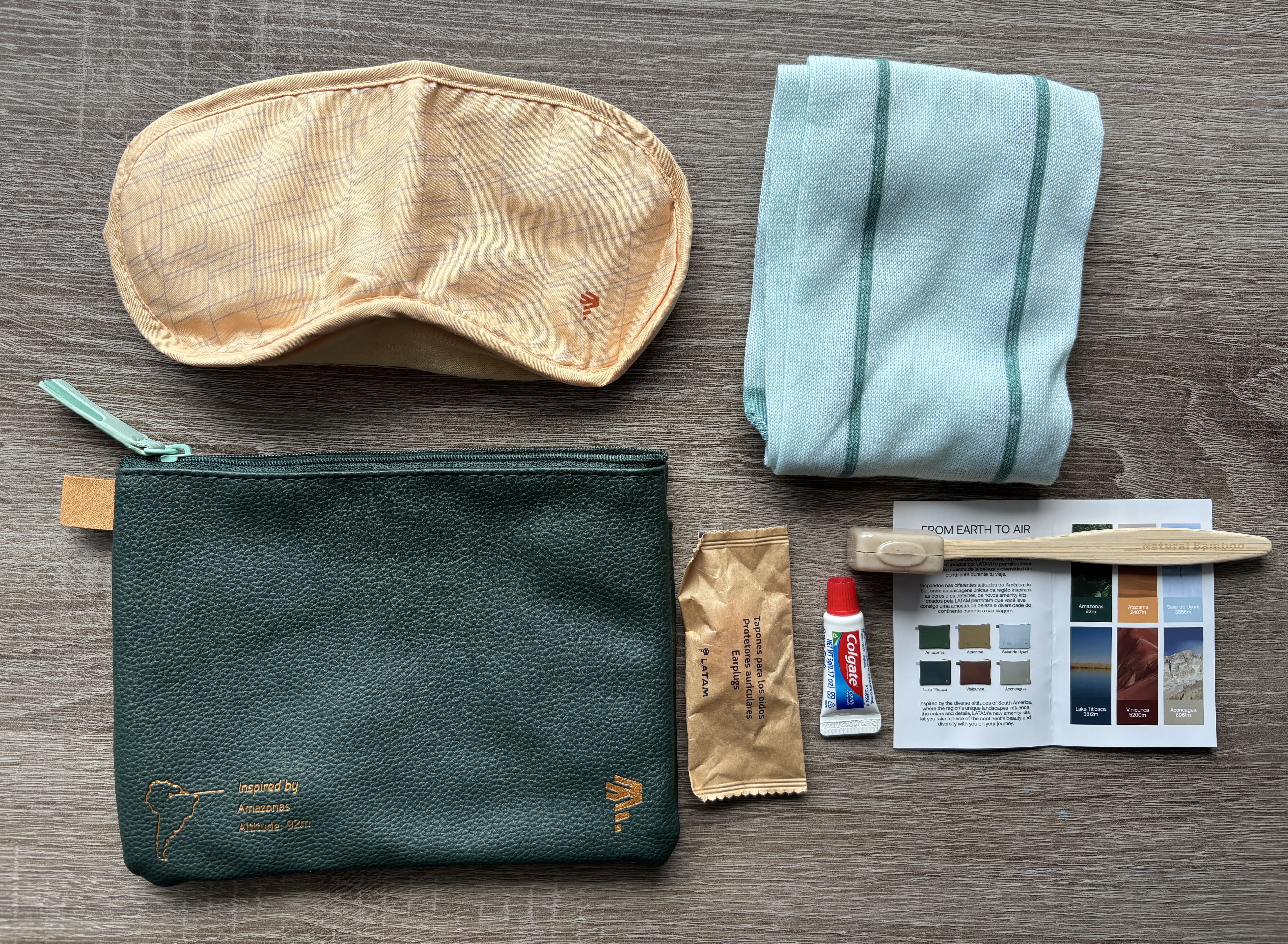 Green LATAM amenity kit bag alongside contents: yellow eyemask, green socks, toothbrush, toothpaste, earplugs, and small booklet explaining the amenity kit design.