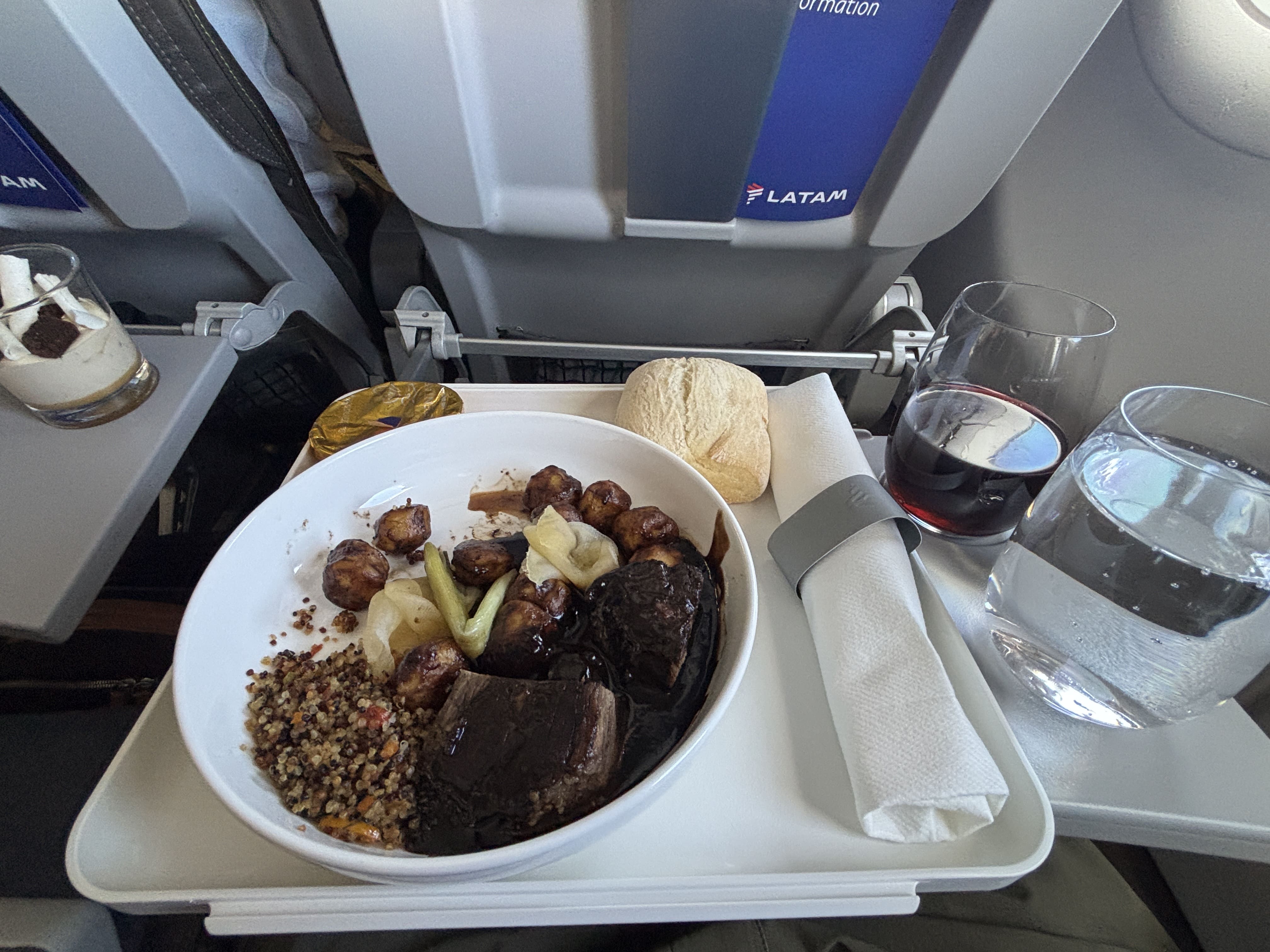 Meal tray with beef, quinoa, and gnocchi, a dessert cup to the left and glasses of red wine and sparkling water to the right.