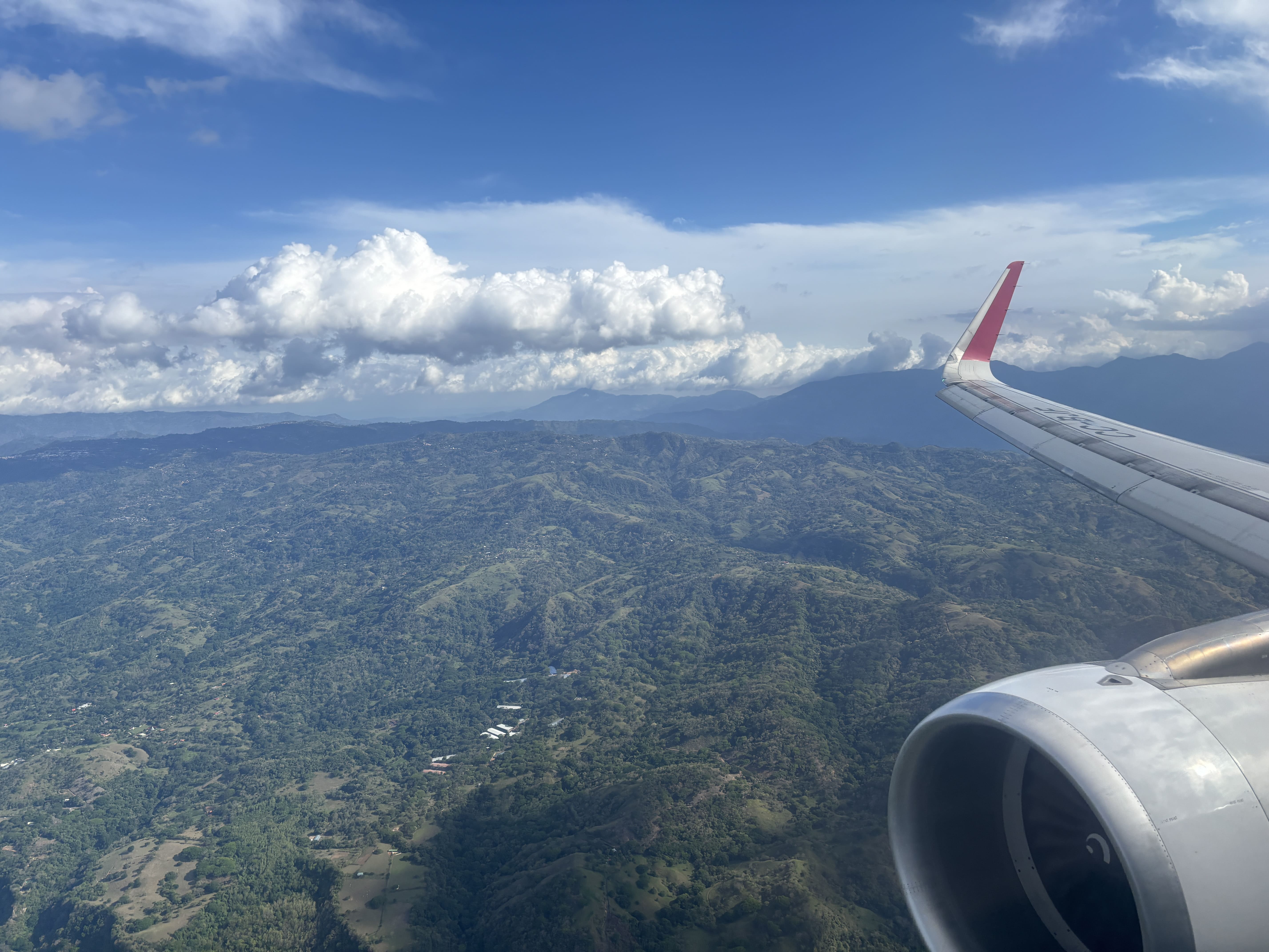 View from an airplane window,showing part of an engine, red wing tip, and green hills.