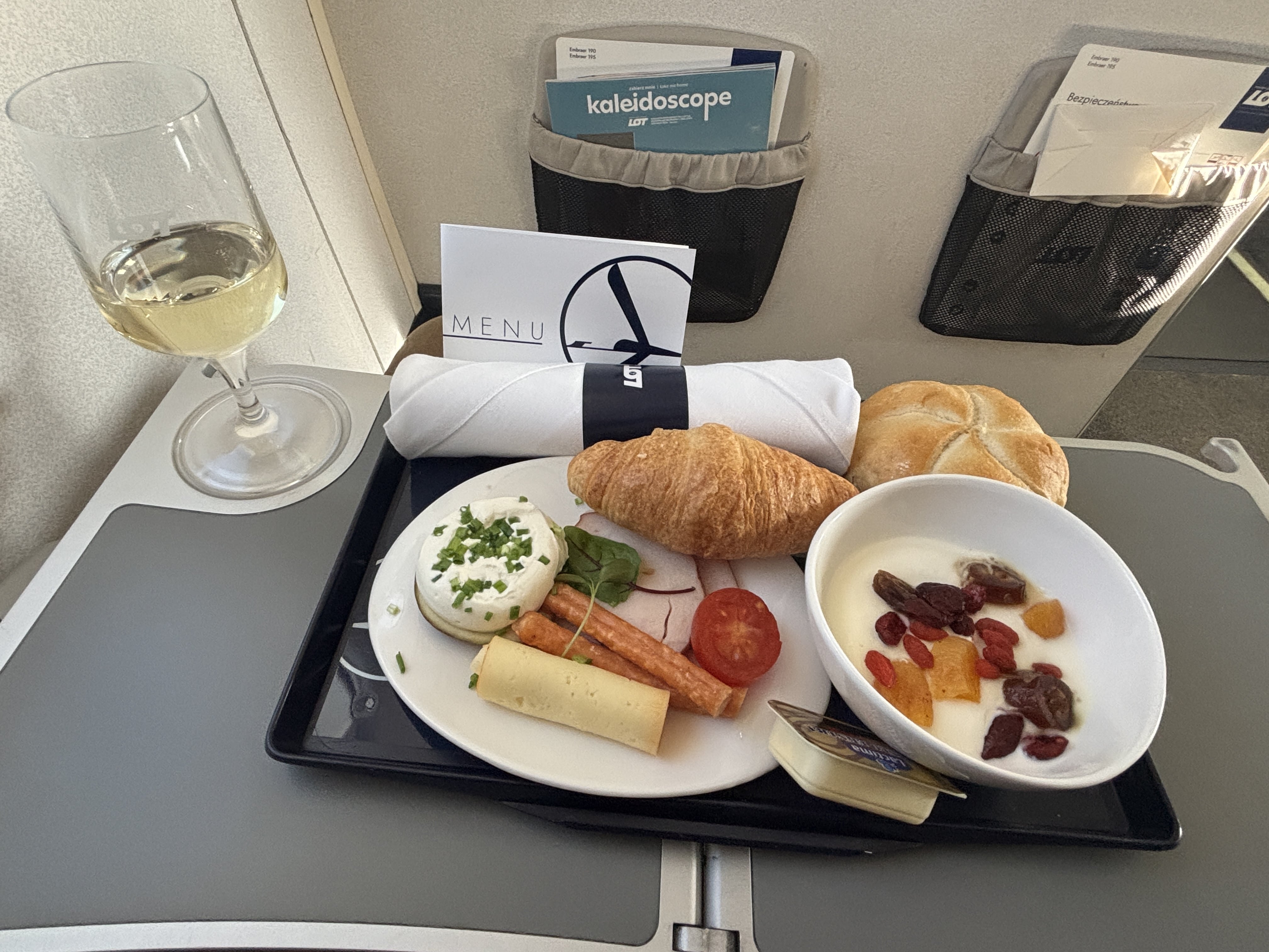 Stemmed glass of sparkling wine resting on an airplane tray table next to a tray holding breakfast: sliced cheeses and meats, yoghurt with candied fruits, and a fig slice with cream cheese, along with croissant and bread roll.