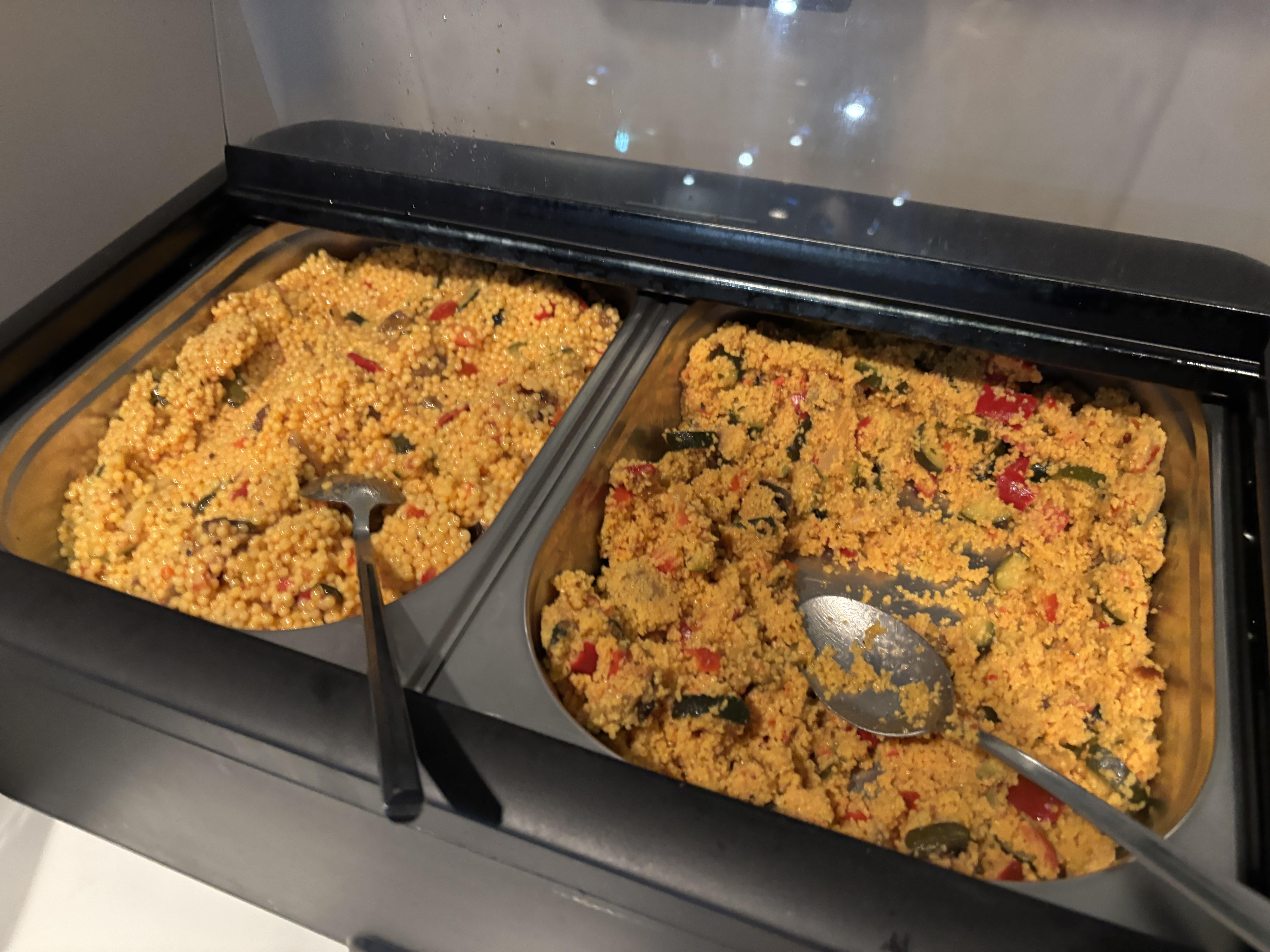 Light-orange coloured couscous with sliced vegetables in side-by-side serving trays, metal spoons protruding from both.