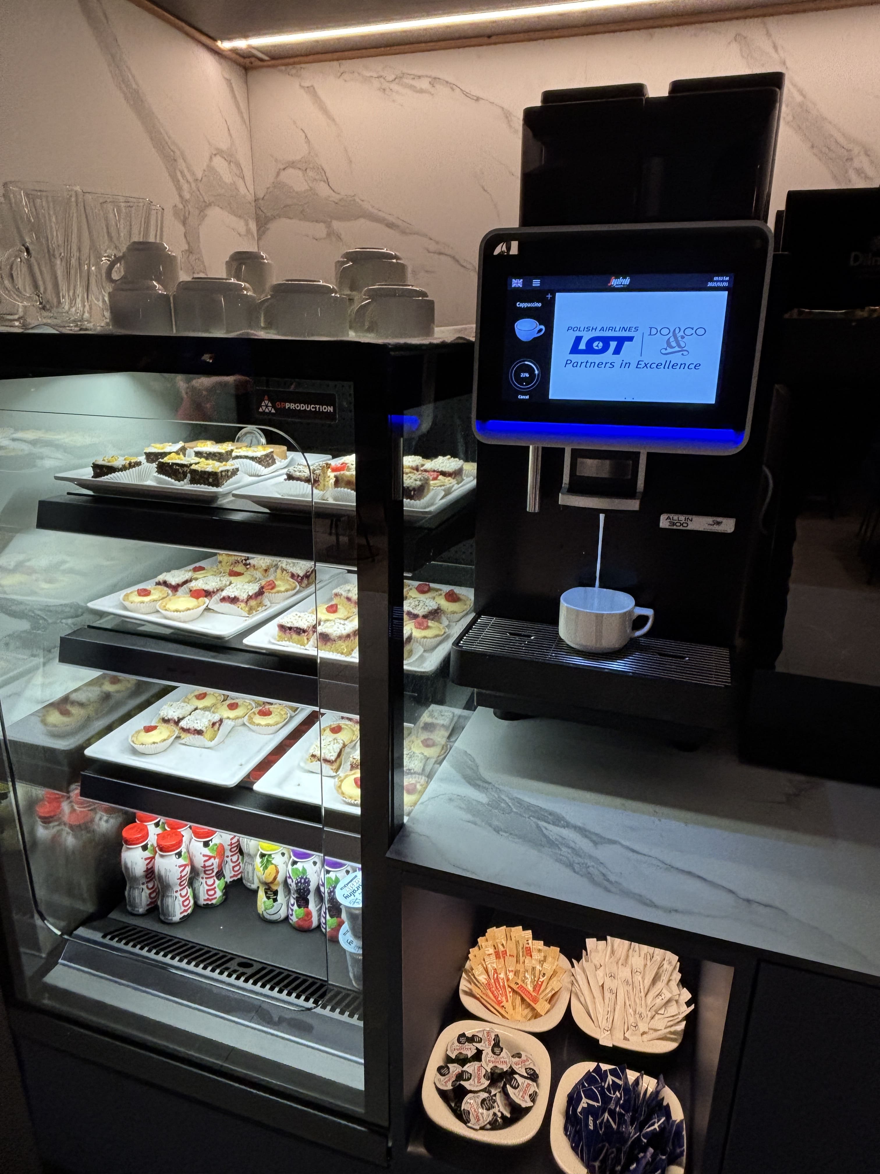 Glass display case containing assorted pastries along with yoghurt-based drinks, next to a faux marble counter on which sits an automated espresso machine.