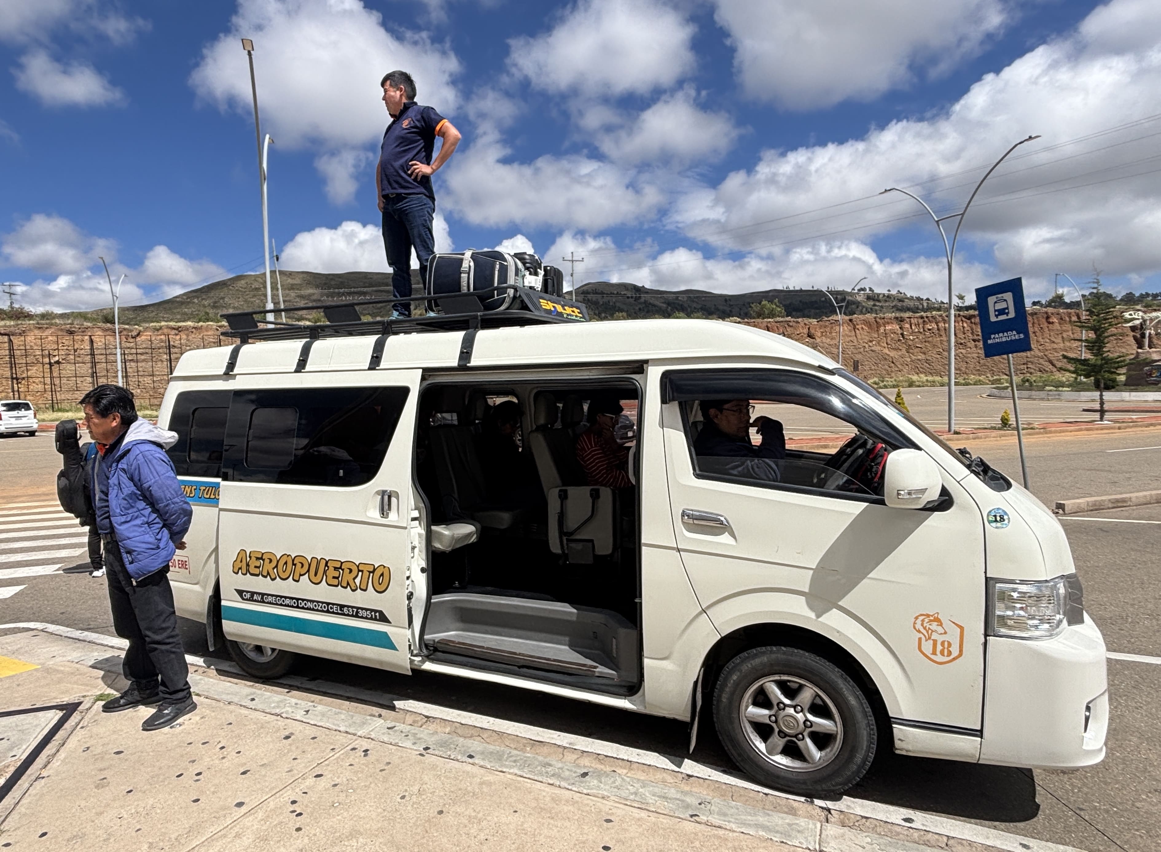 White passenger van parked at Alcantarí Airport, showing a man standing on top loading luggage.