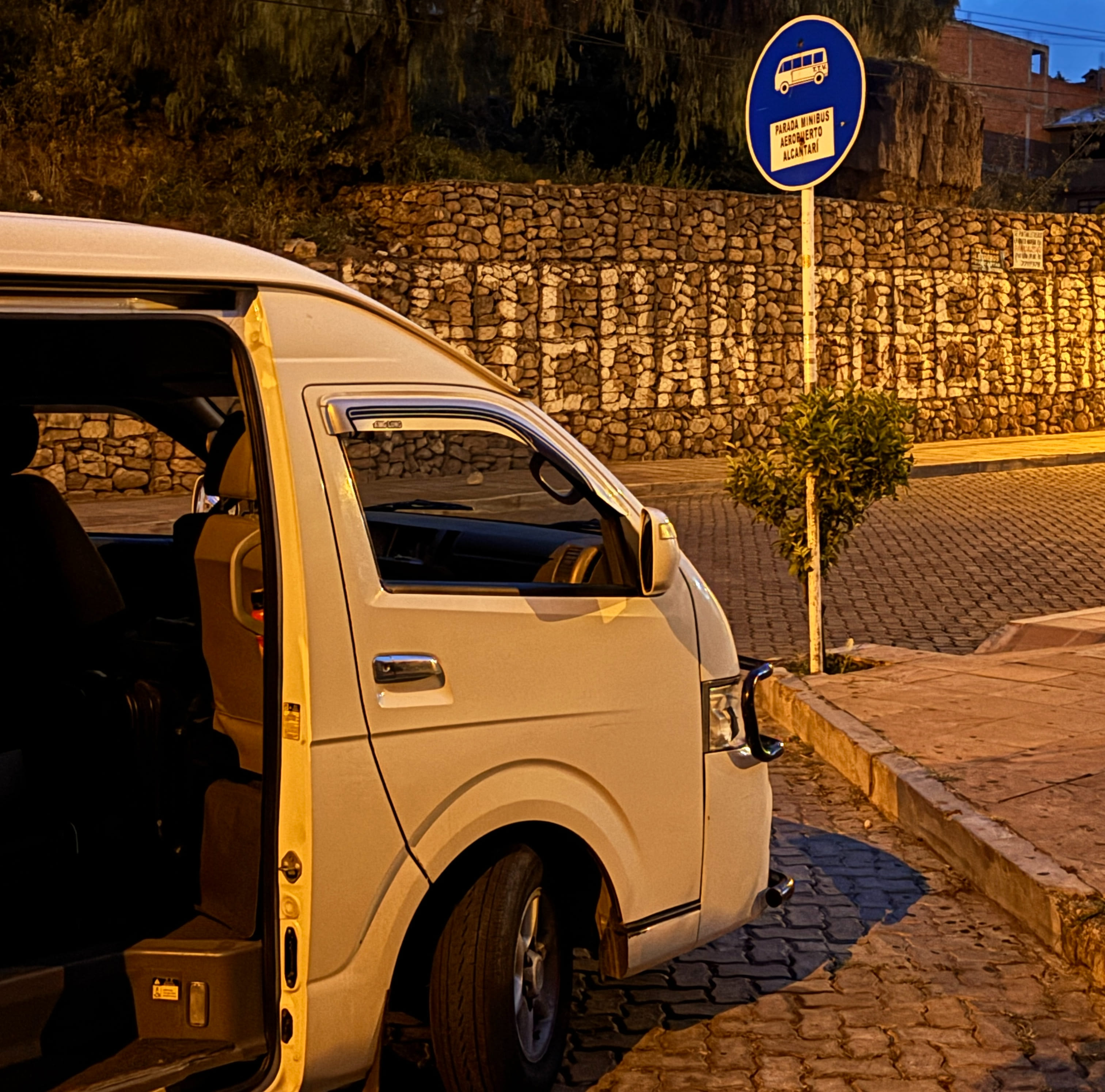Front half of a white passenger van waiting next to a blue bus stop sign.