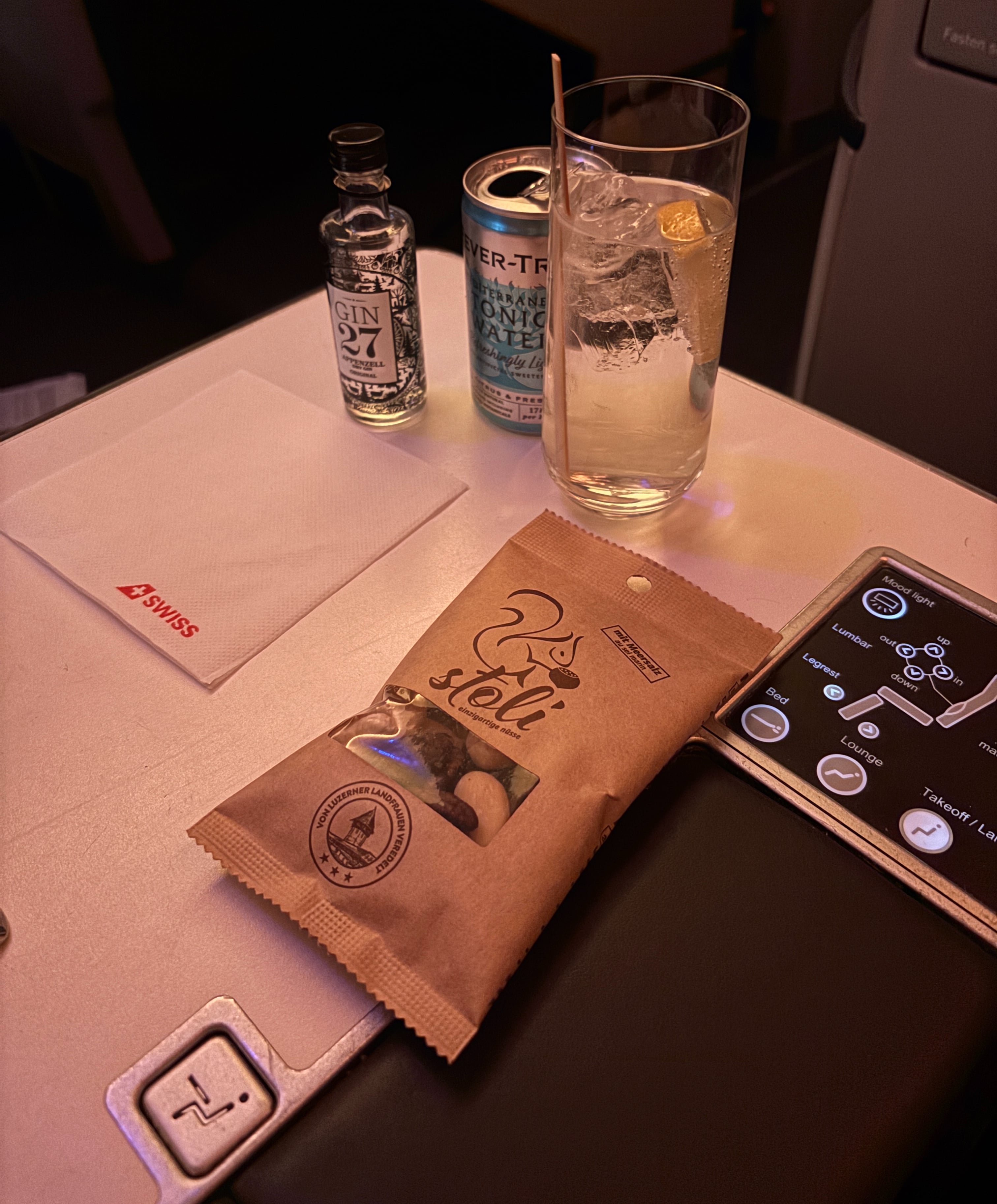 From left to right: small glass bottle of Gin 27, can of Fever-Tree tonic water, gin and tonic in a glass with ice and a lemon slice, and a brown paper bag of mixed nuts.