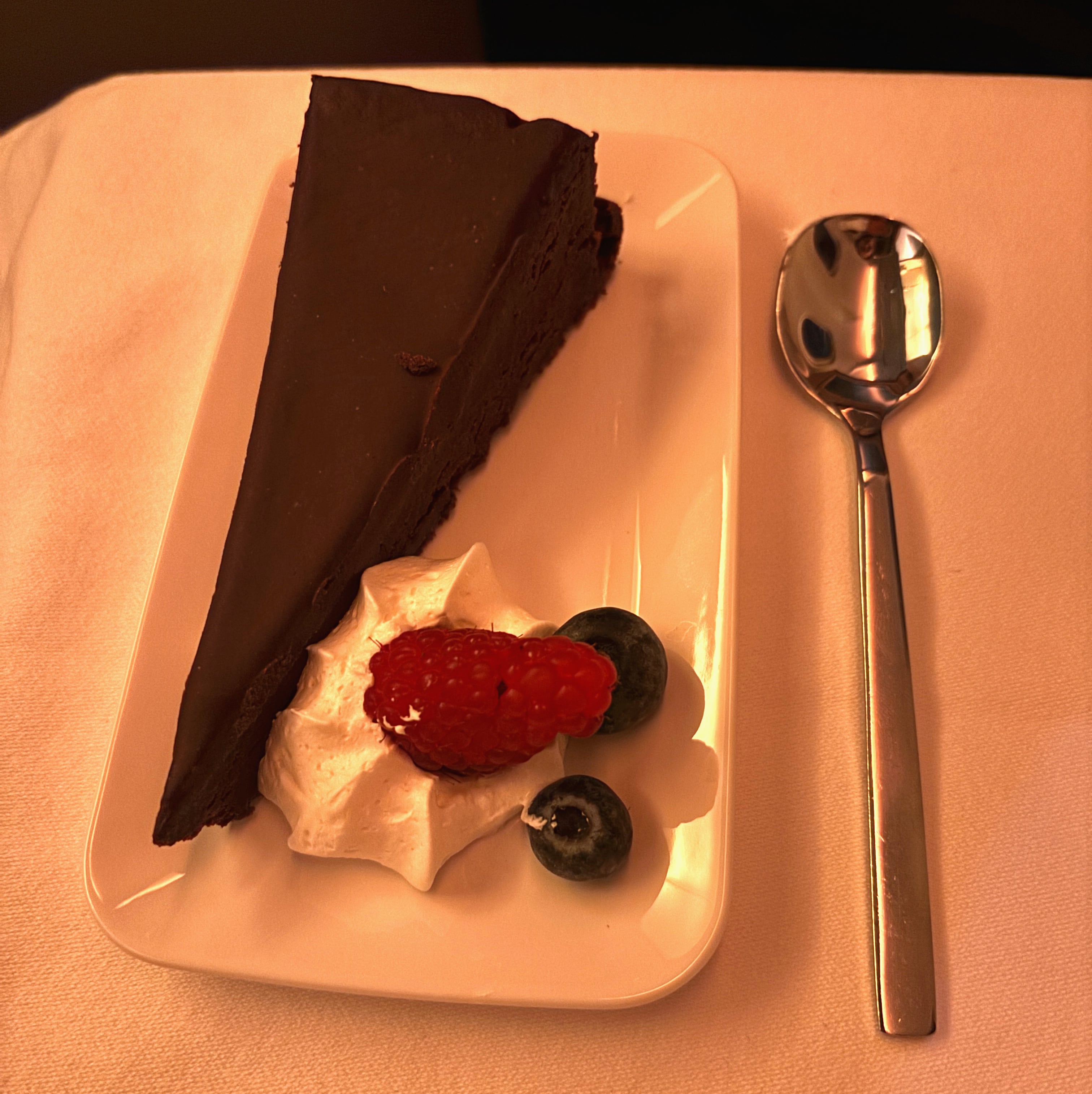 Slice of chocolate cake served on white plate with berries and whipped cream, metal spoon on the right side.