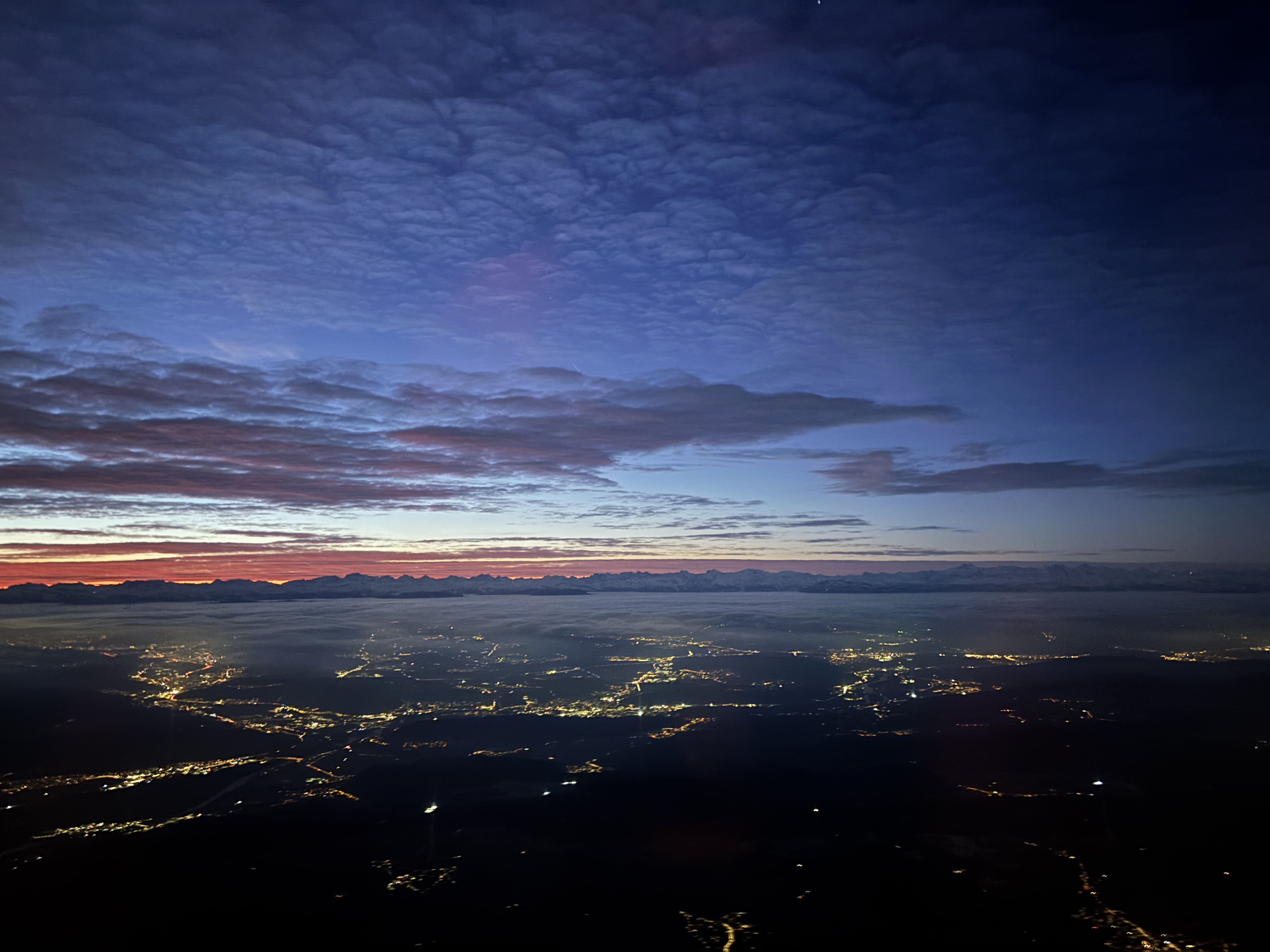 Sliver of pinkish-orange seen in the distance over a smattering of street lights seen from the air beneath a cloudly sky as the sun begins to rise.