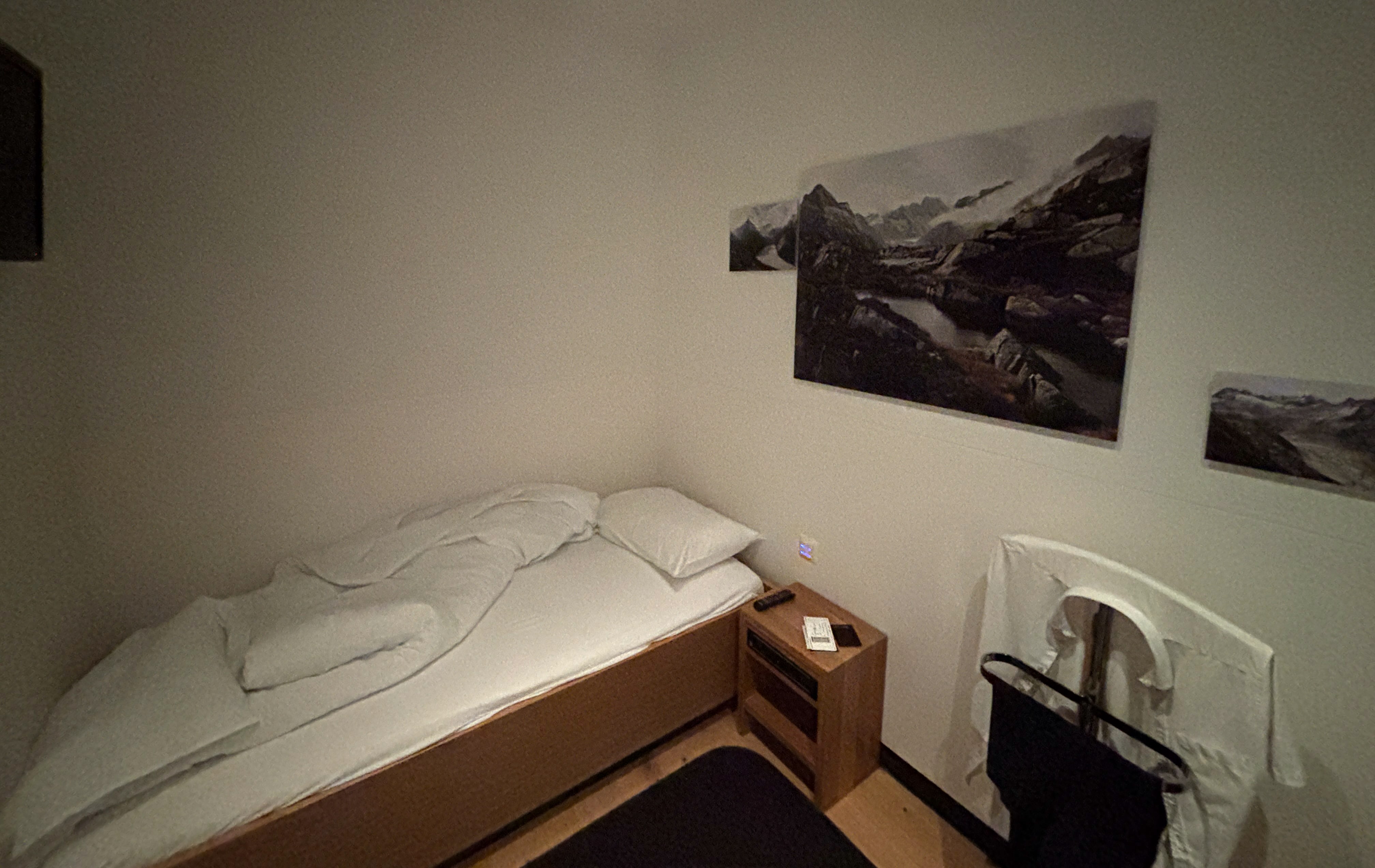 White bedroom with black-and-white digital prints of mountains on one wall, small bed next to furthest wall, corner of television facing bed, and a garment stand with hanging shirt.