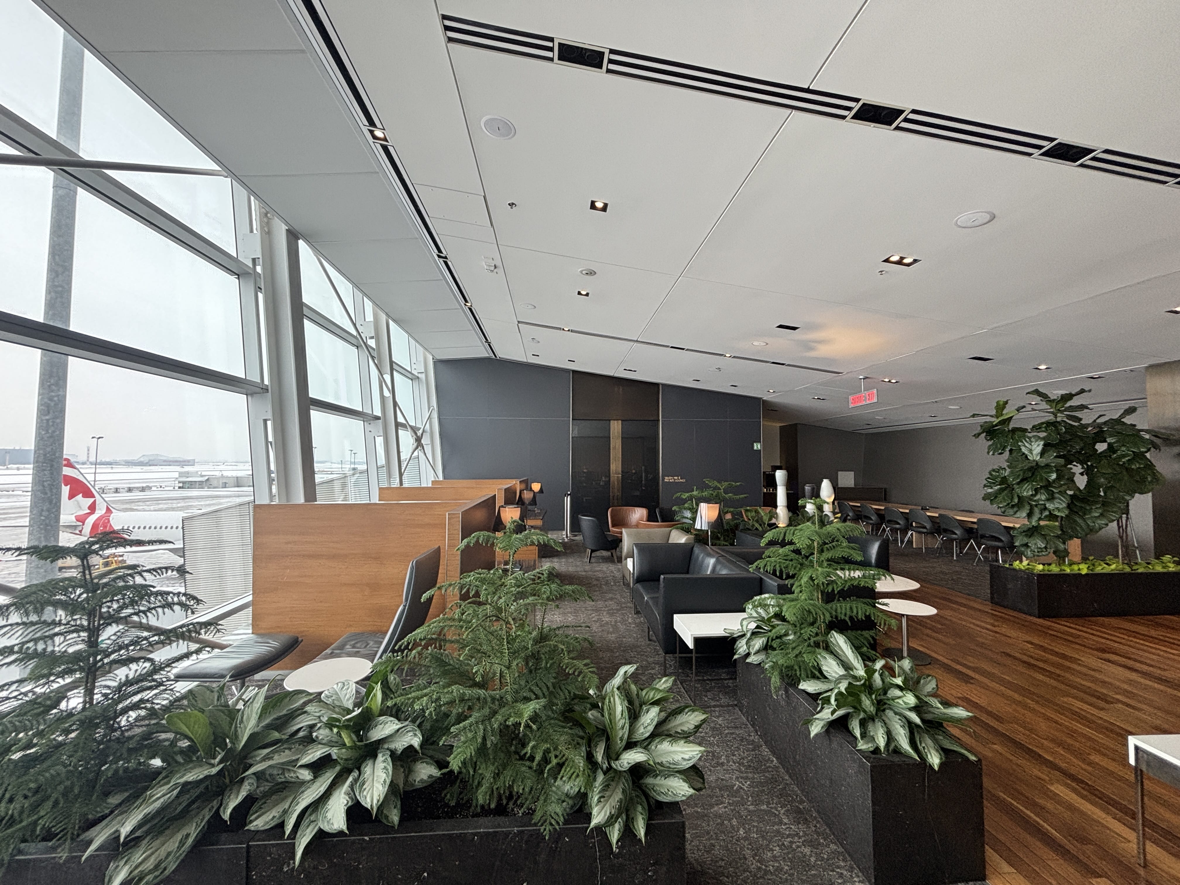 Black lounge chairs with brown wood dividers arranged facing a floor-to-ceiling window, with the tailfin of an Air Canada Rouge jet visible.