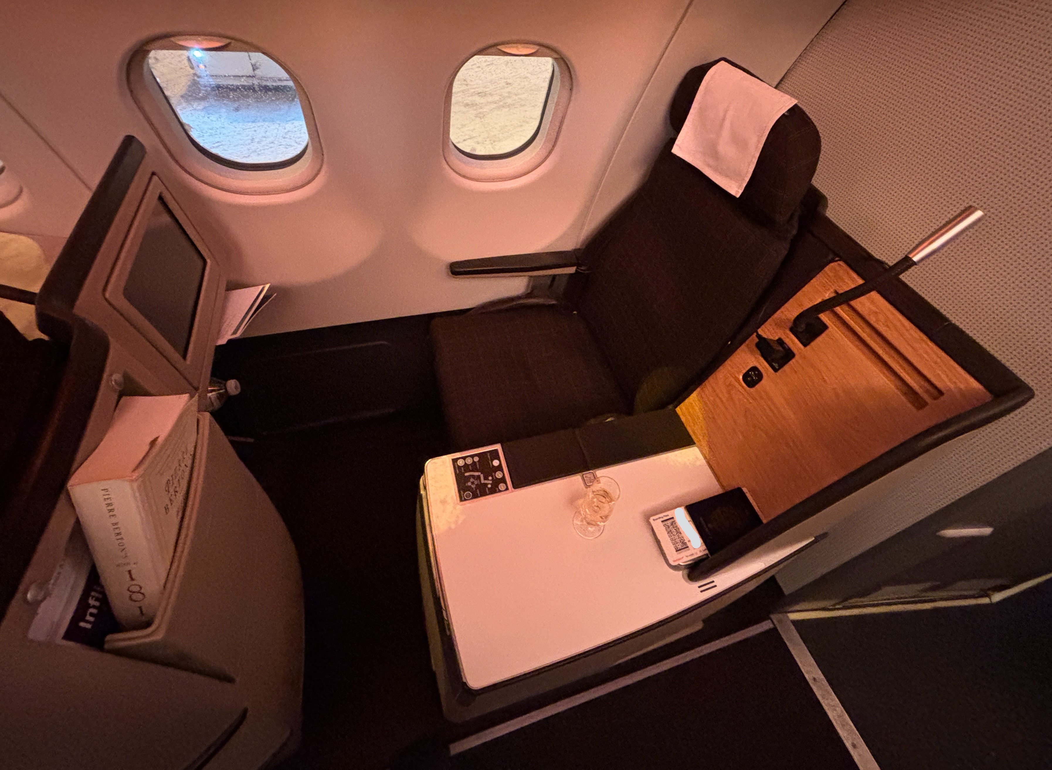 Brown seat with white headrest next to two airplane windows, white table to the seat's left with wooden accent and reading light above.  A small screen faces the seat.
