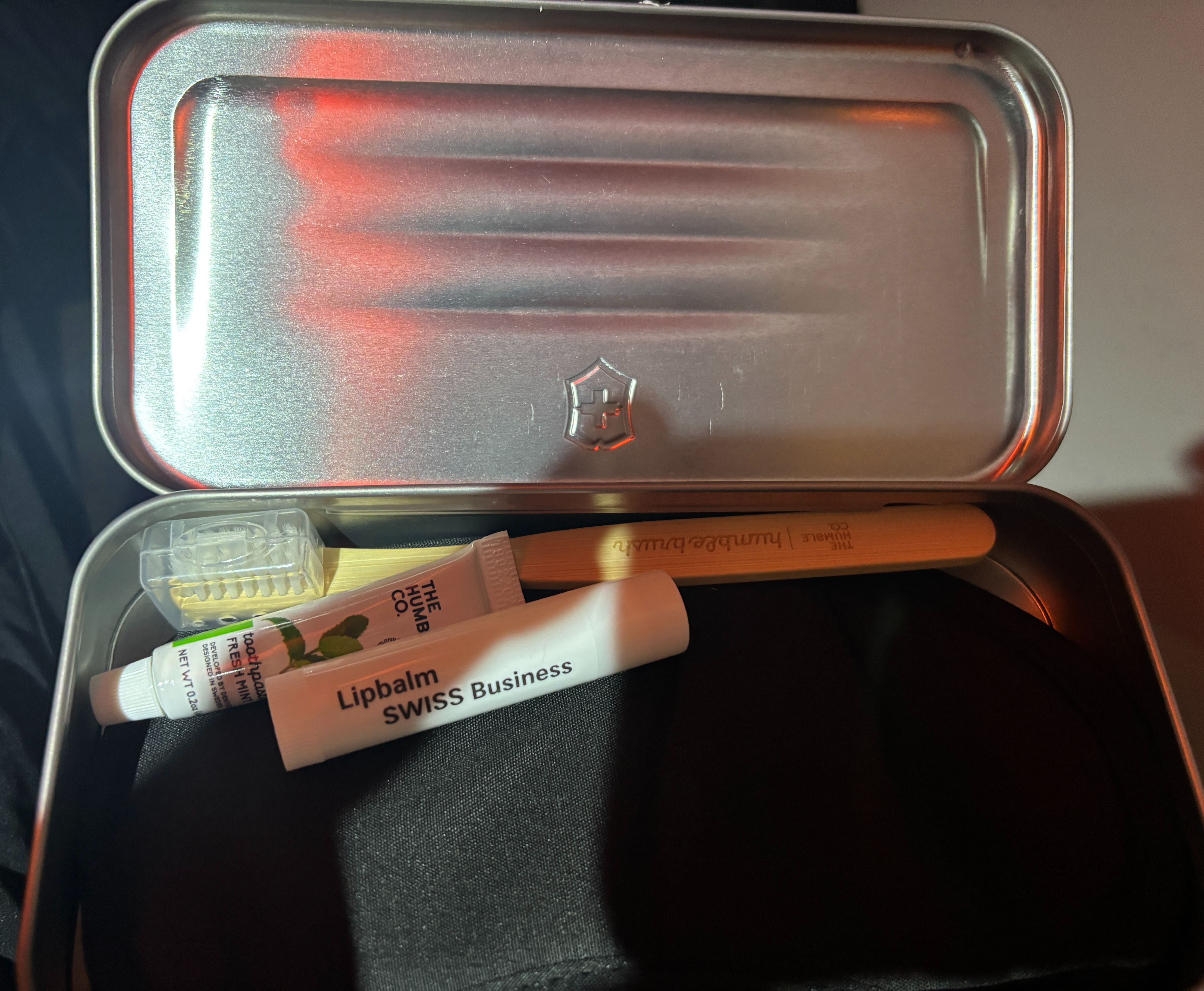 Opened metallic box showing small bamboo toothbrush, black eye mask, and two white tubes of lip balm and toothpaste.