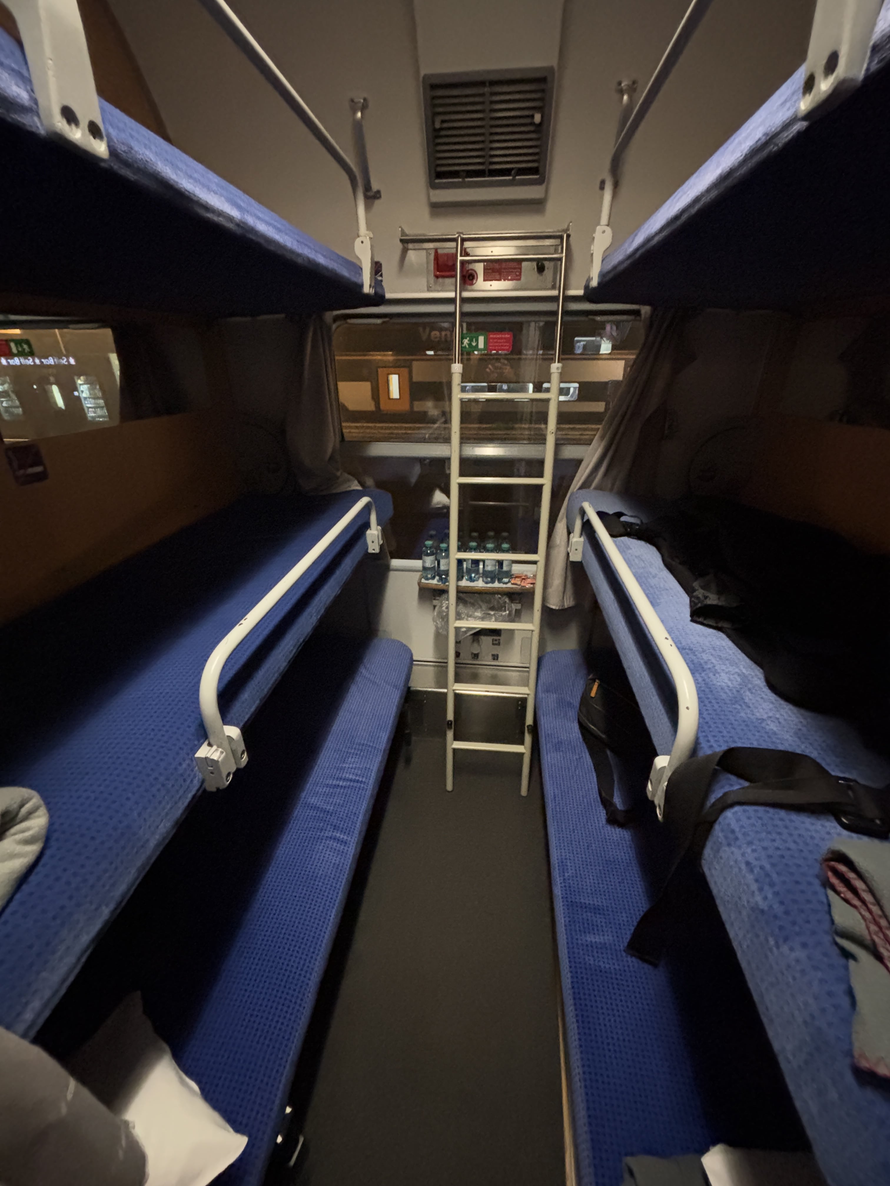 Interior view of couchette cabin, showing three blue mattresses stacked vertically on either side of a small floor, with ladder in the middle against an exterior-facing window.