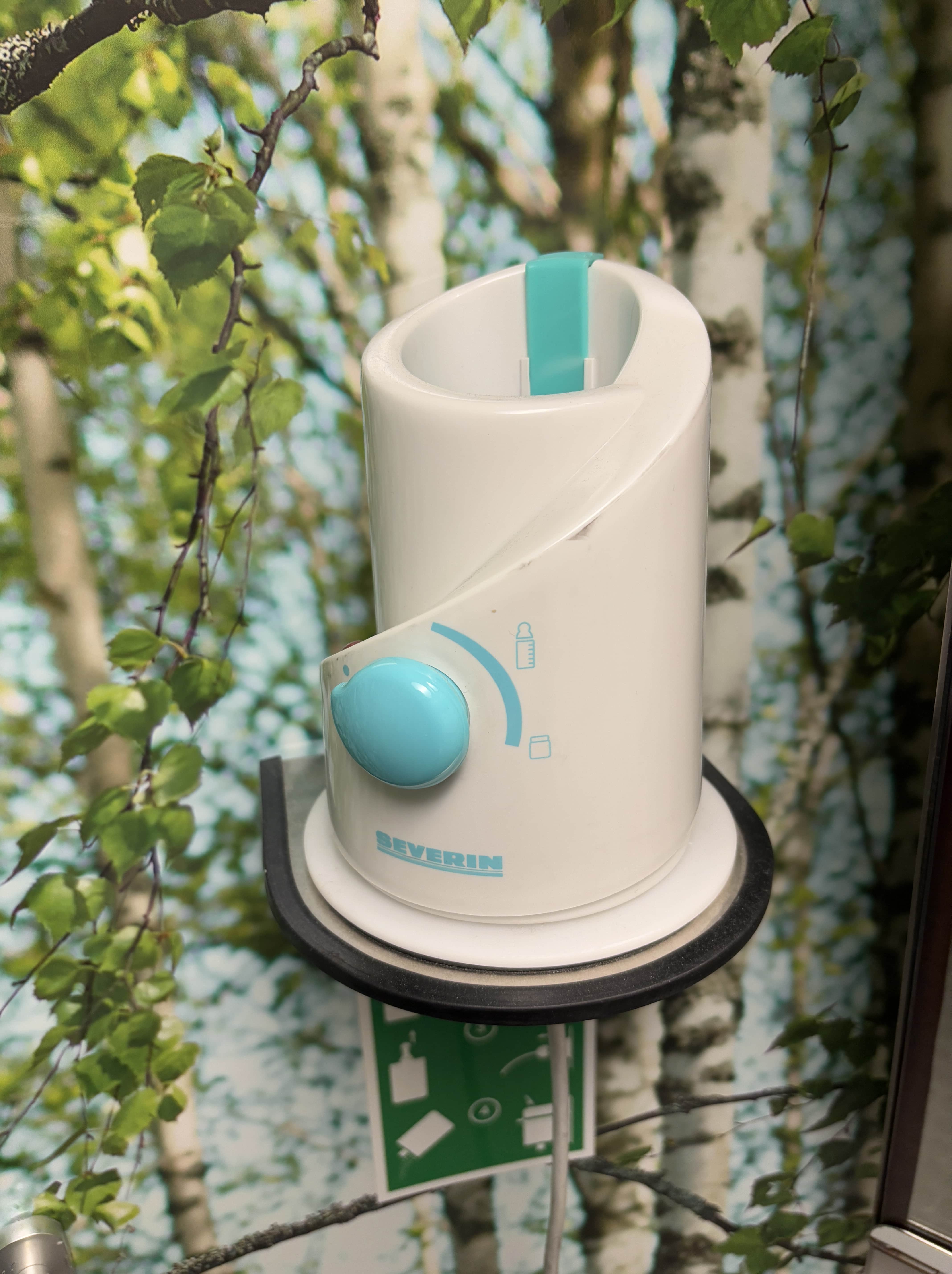 Baby bottle warmer affixed to lavatory wall, with trees on the wallpaper.