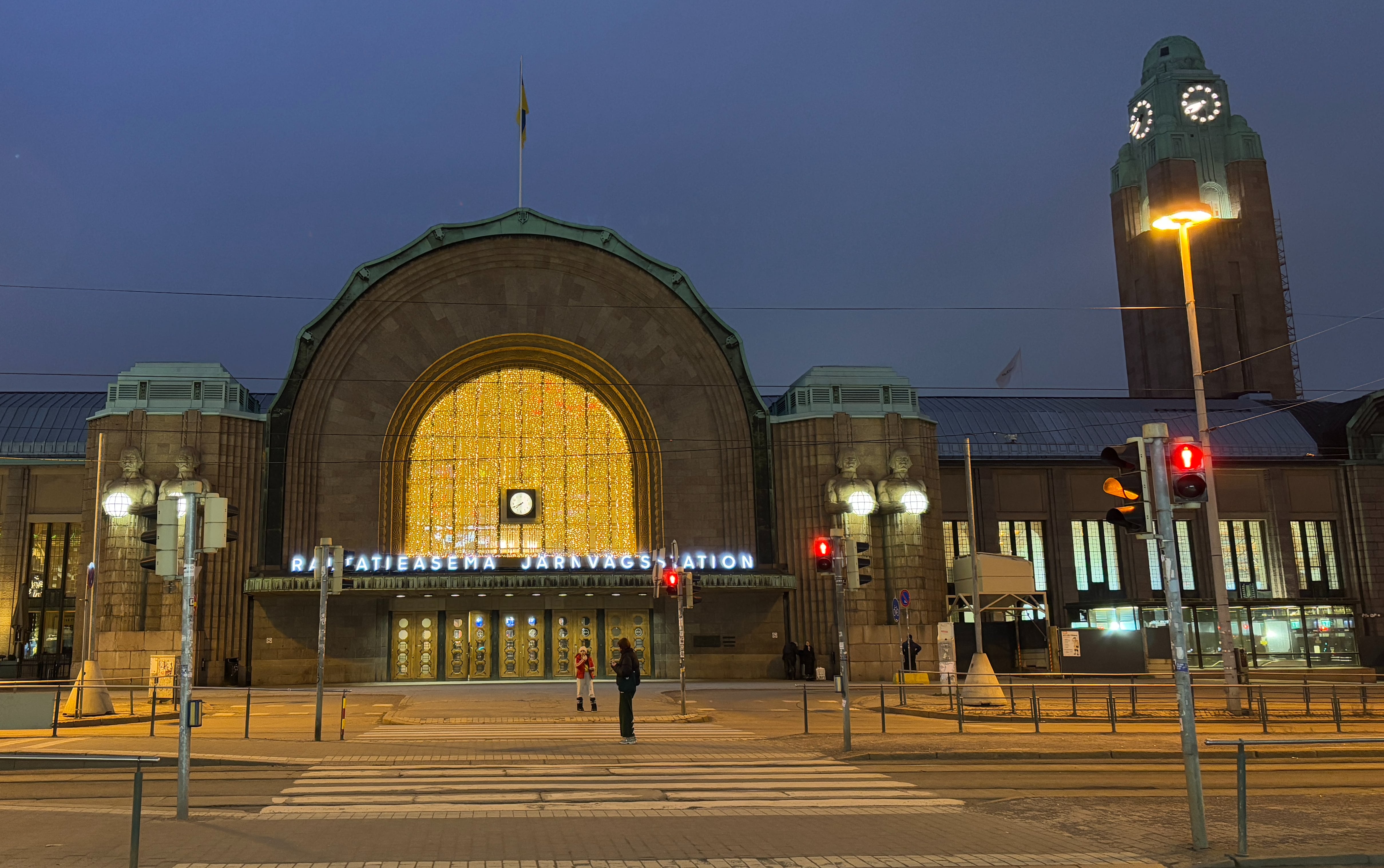 Helsinki train station seen from pedestrian crossing.  Dark early-morning sky is seen behind the station's well-lit entrance and clock tower.