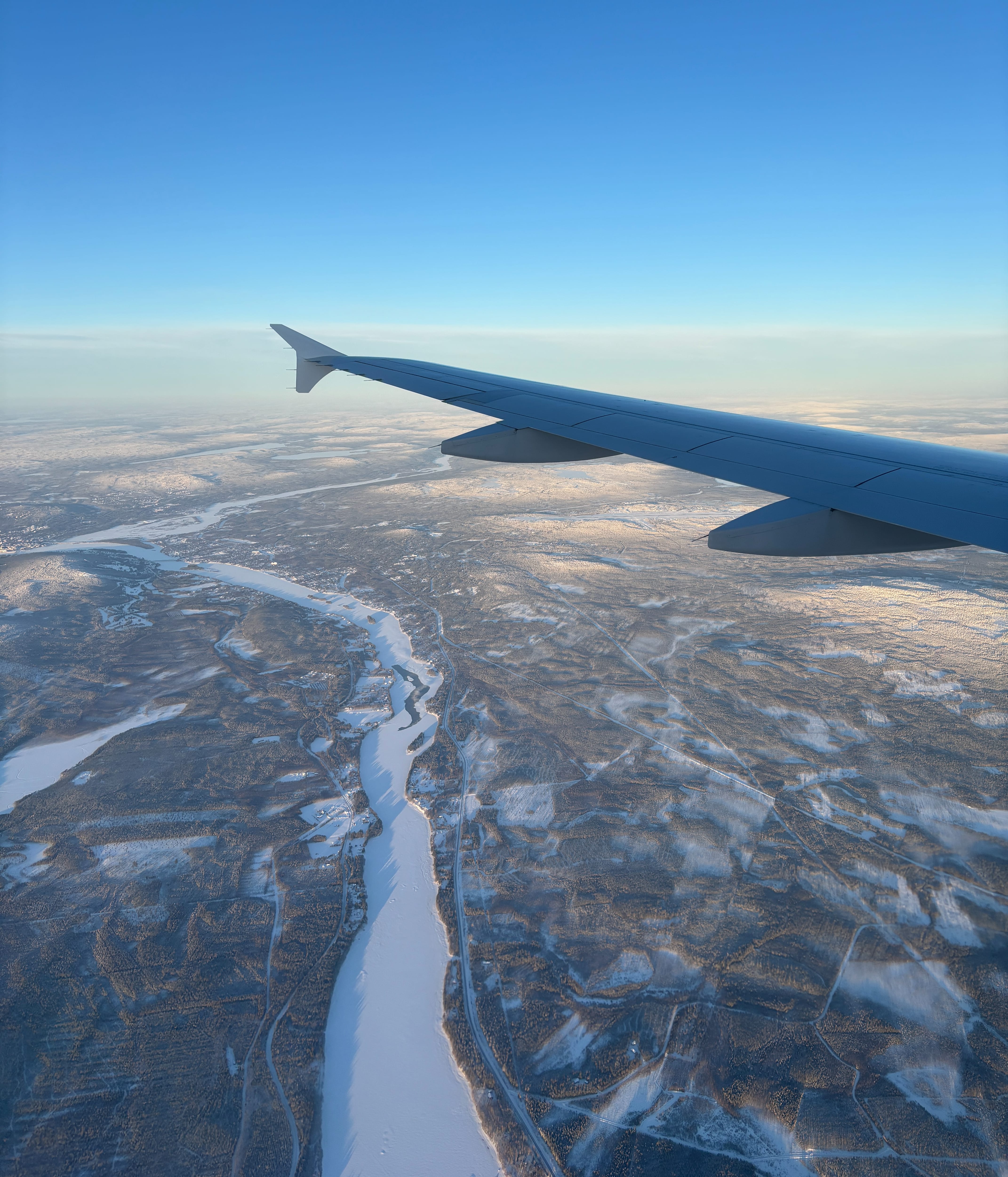 Clear blue sky with frozen, snow-covered river below, seen from plane window with wing visible covering the horizon.