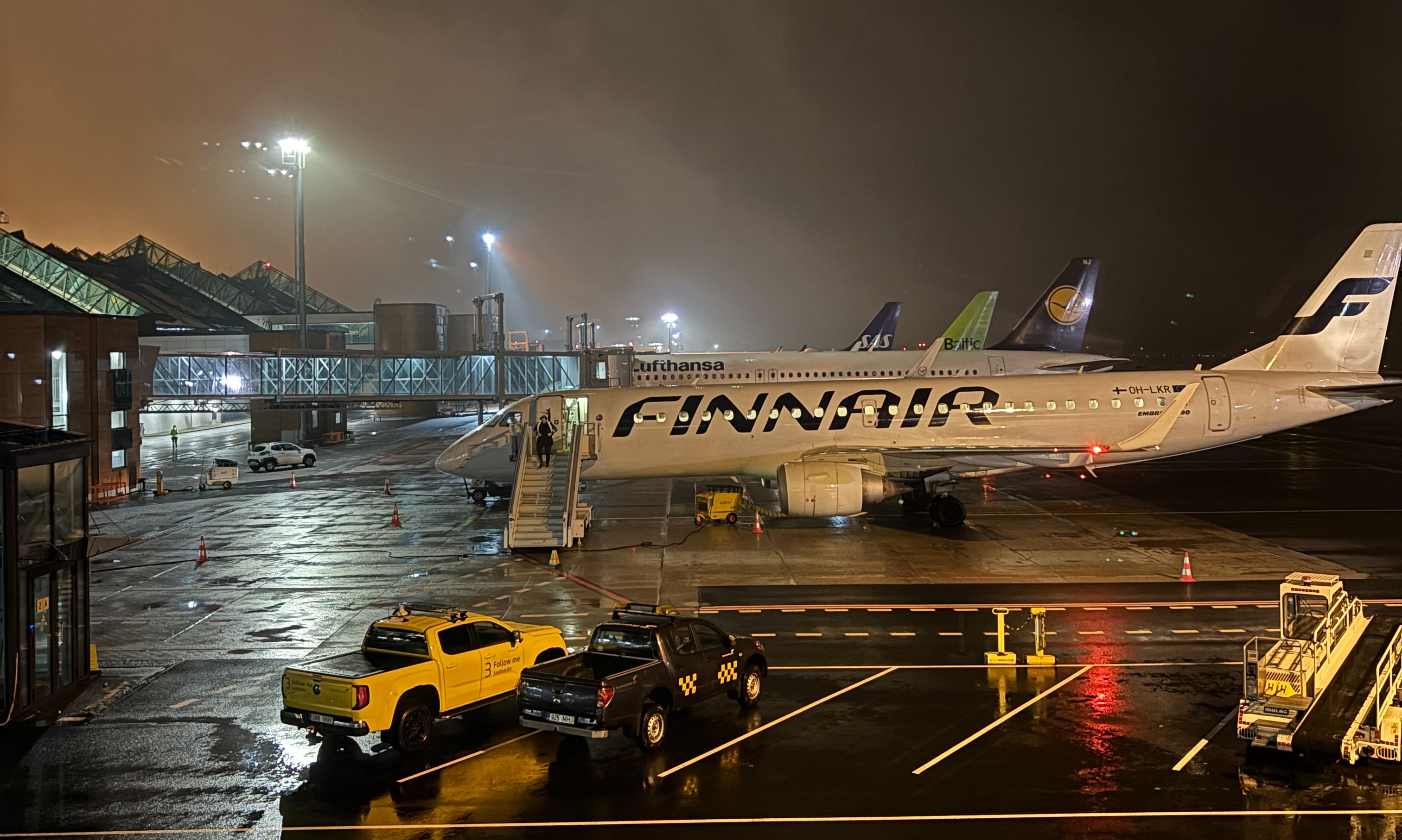 Finnair regional jet sitting on the apron connected to airstairs in the early morning, tailfins of Lufthansa, airBaltic, and SAS planes visible in the background.