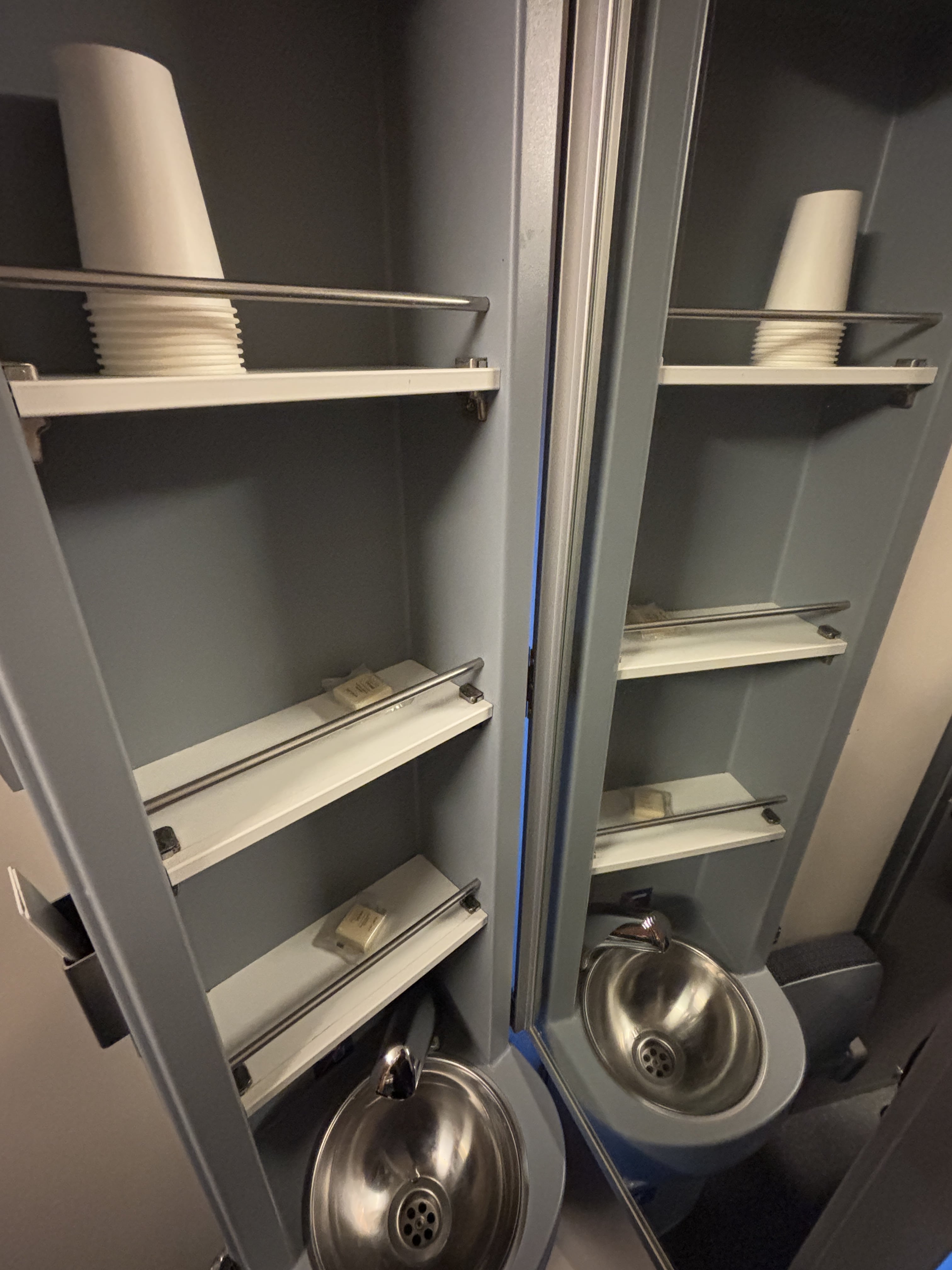 Small sink below shelves containing paper cups and two small bars of soap, with mirror contained in folding door which closes to conceal the washbasin.