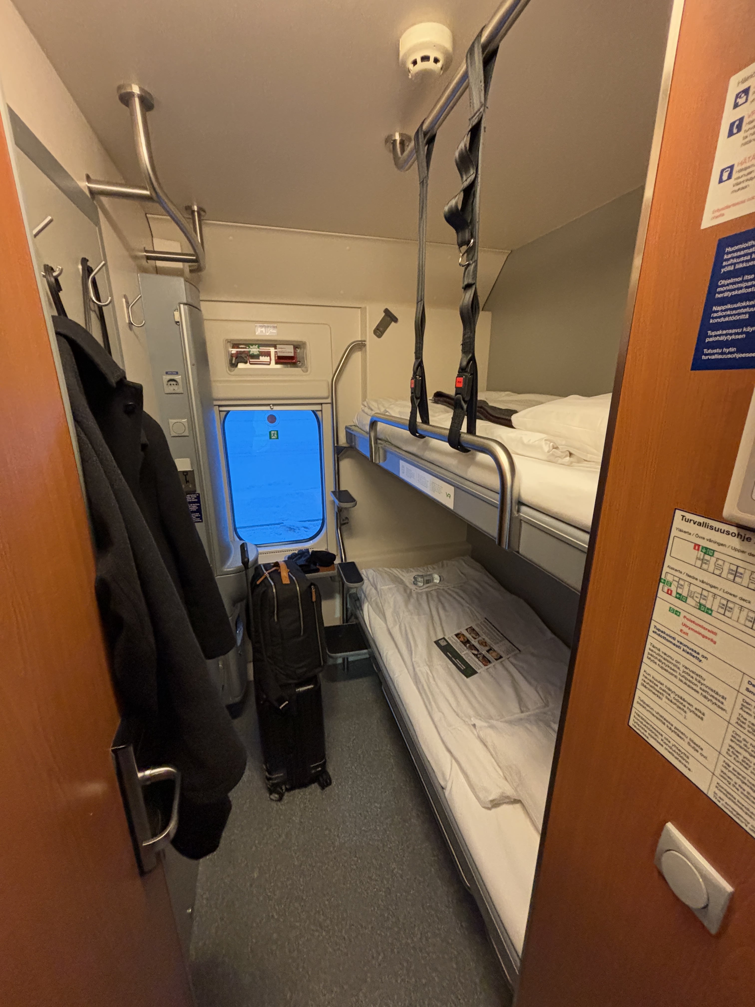 Floor-to-ceiling photo of dual-berth cabin aboard VR night train.  A winter jacket hangs from one of four coathooks to the left, small carry-on suitcase is occupying roughly 15% of the floorspace, placed in front of a small exterior-facing window of about the same size as the carry-on.  Two twin beds, one atop the other, are made up with white linens, a bottle of water resting on each.